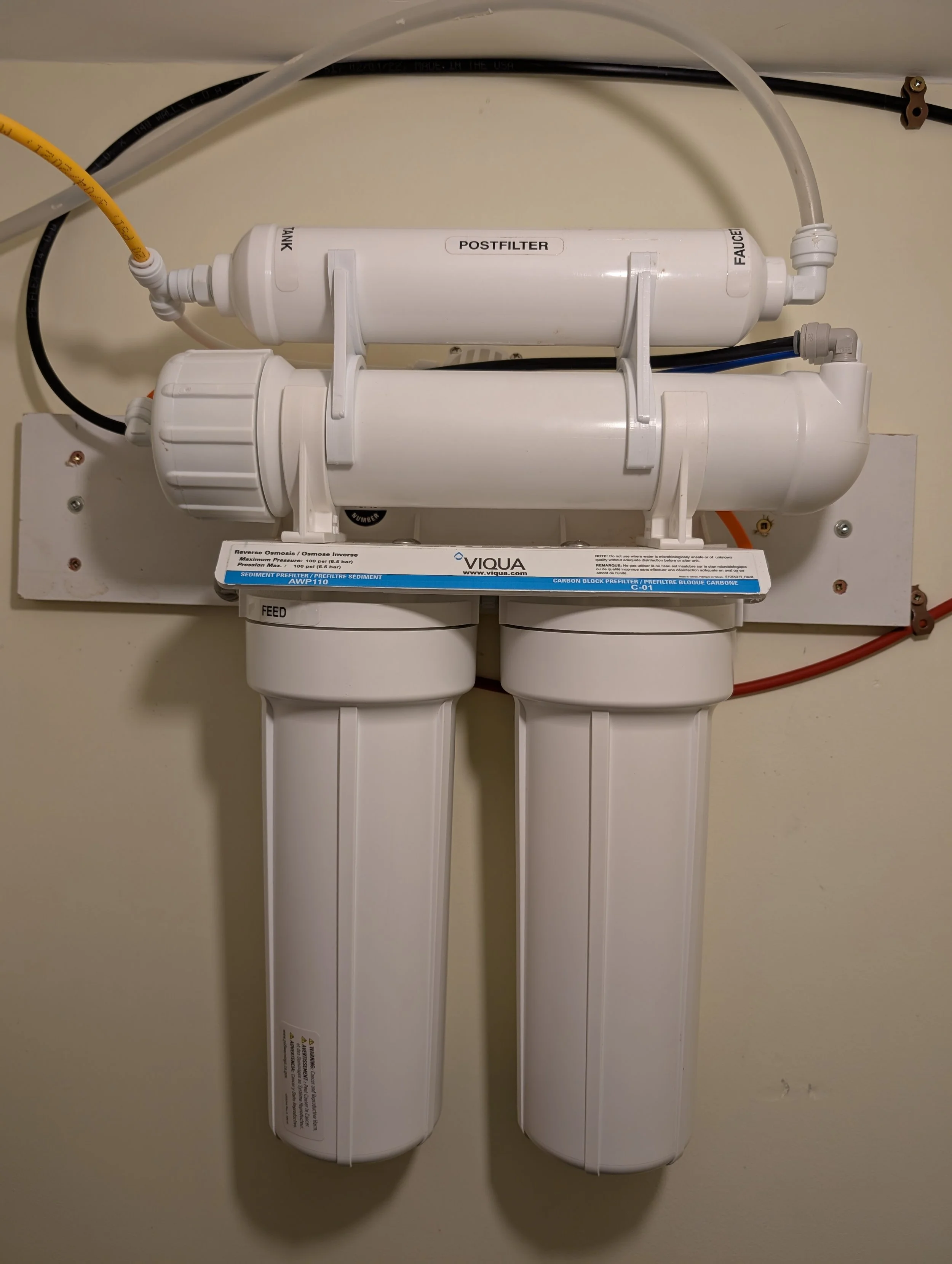 A water filtration system with two white cylindrical filter units mounted vertically on a wall, connected by black and gray tubing, labeled with the brand Viqua and function details.
