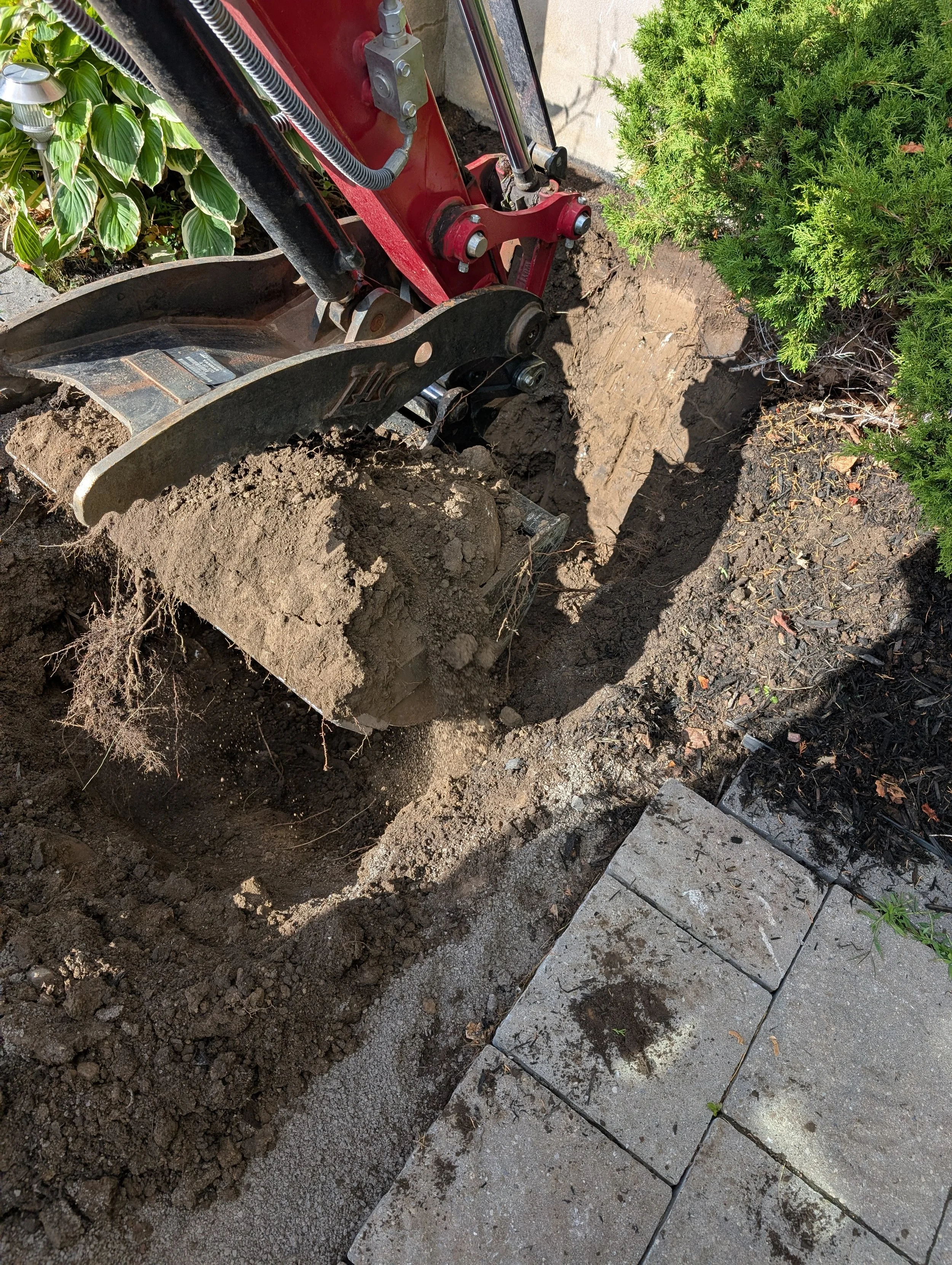 Close-up of a mini excavator digging soil near a paving stone border, with green bushes in the background.
