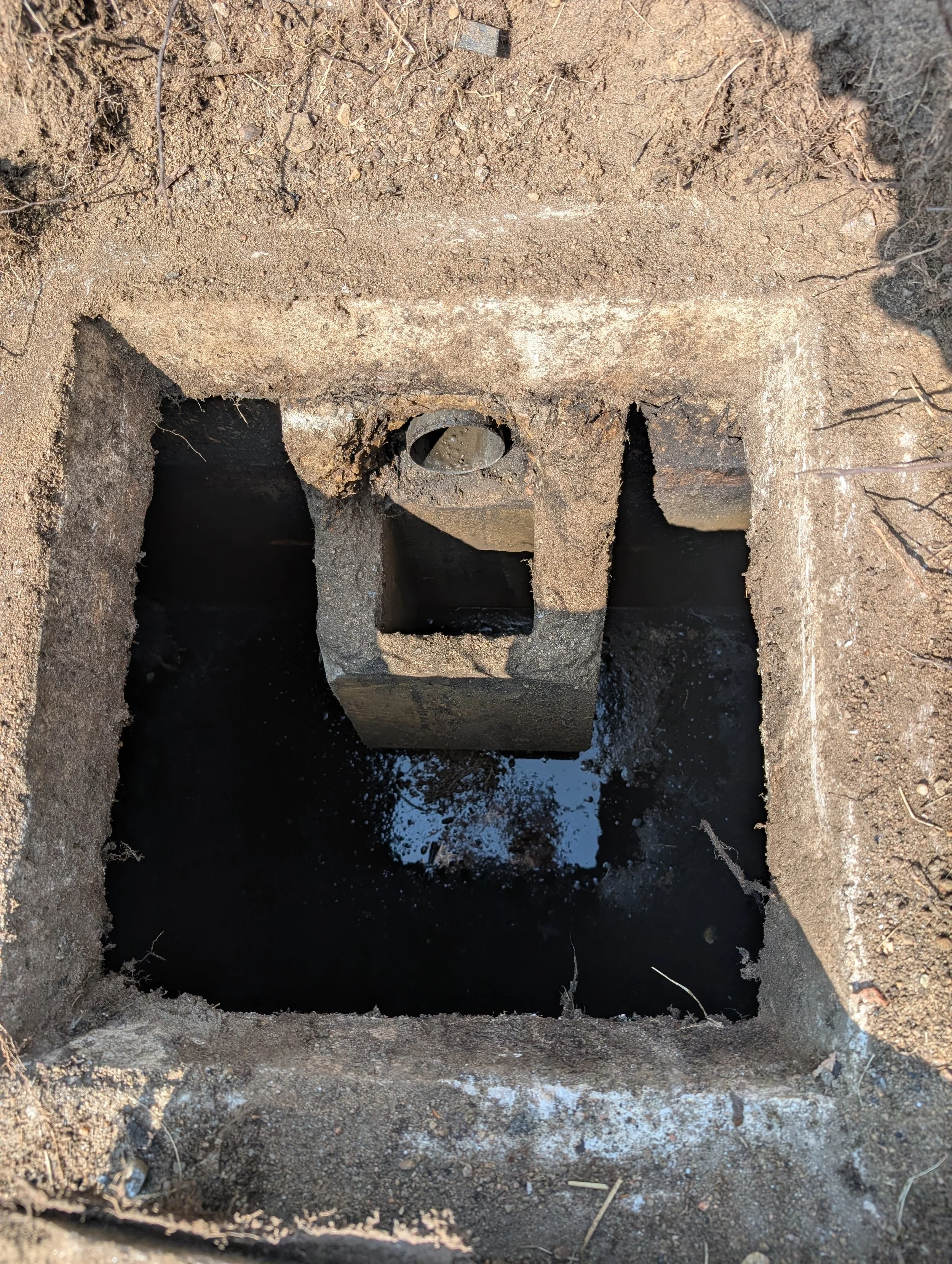 A rectangular opening in the ground with a large stone block at the bottom and an iron pipe protruding from the middle of the block, surrounded by dirt and some roots.