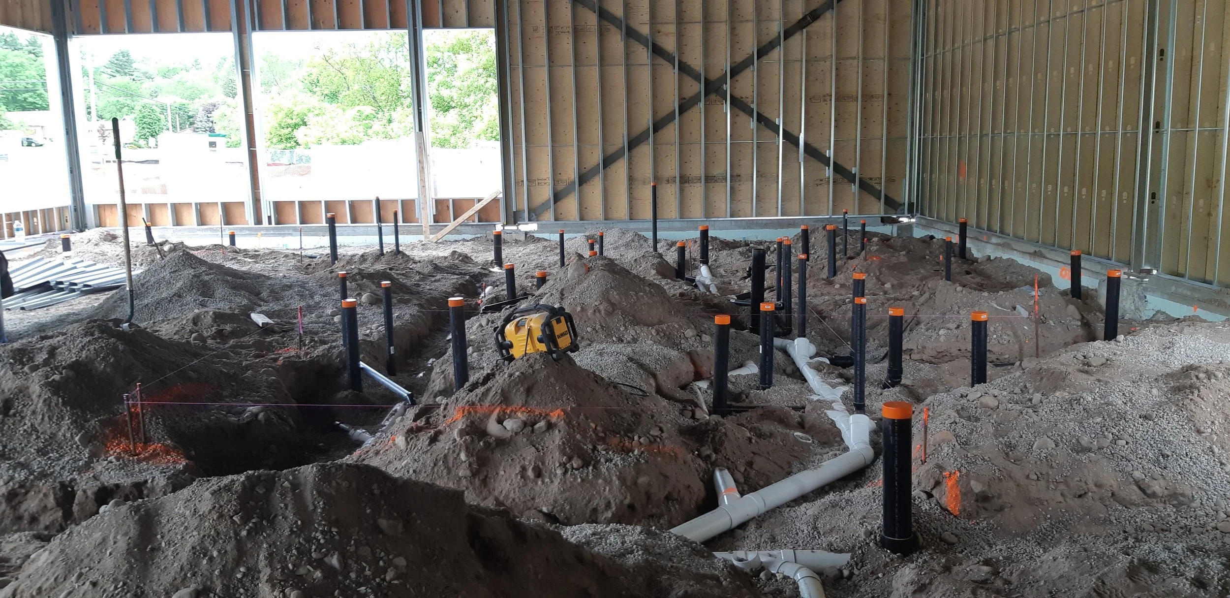 Construction site interior with exposed framing, plumbing pipes, and dirt ground for building foundation.