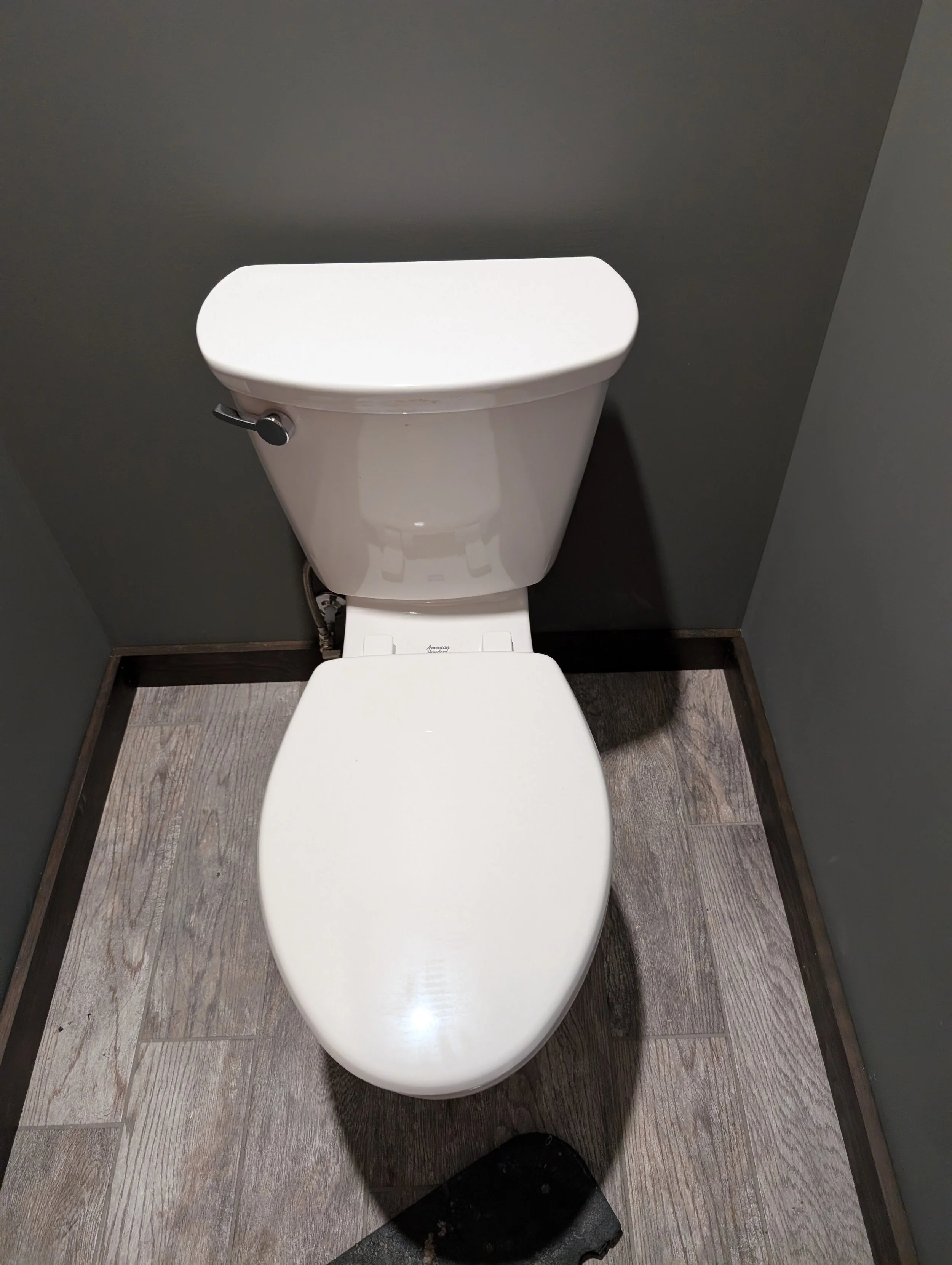 A white toilet with a closed lid, positioned against dark gray walls and gray wood-like floor tiles.