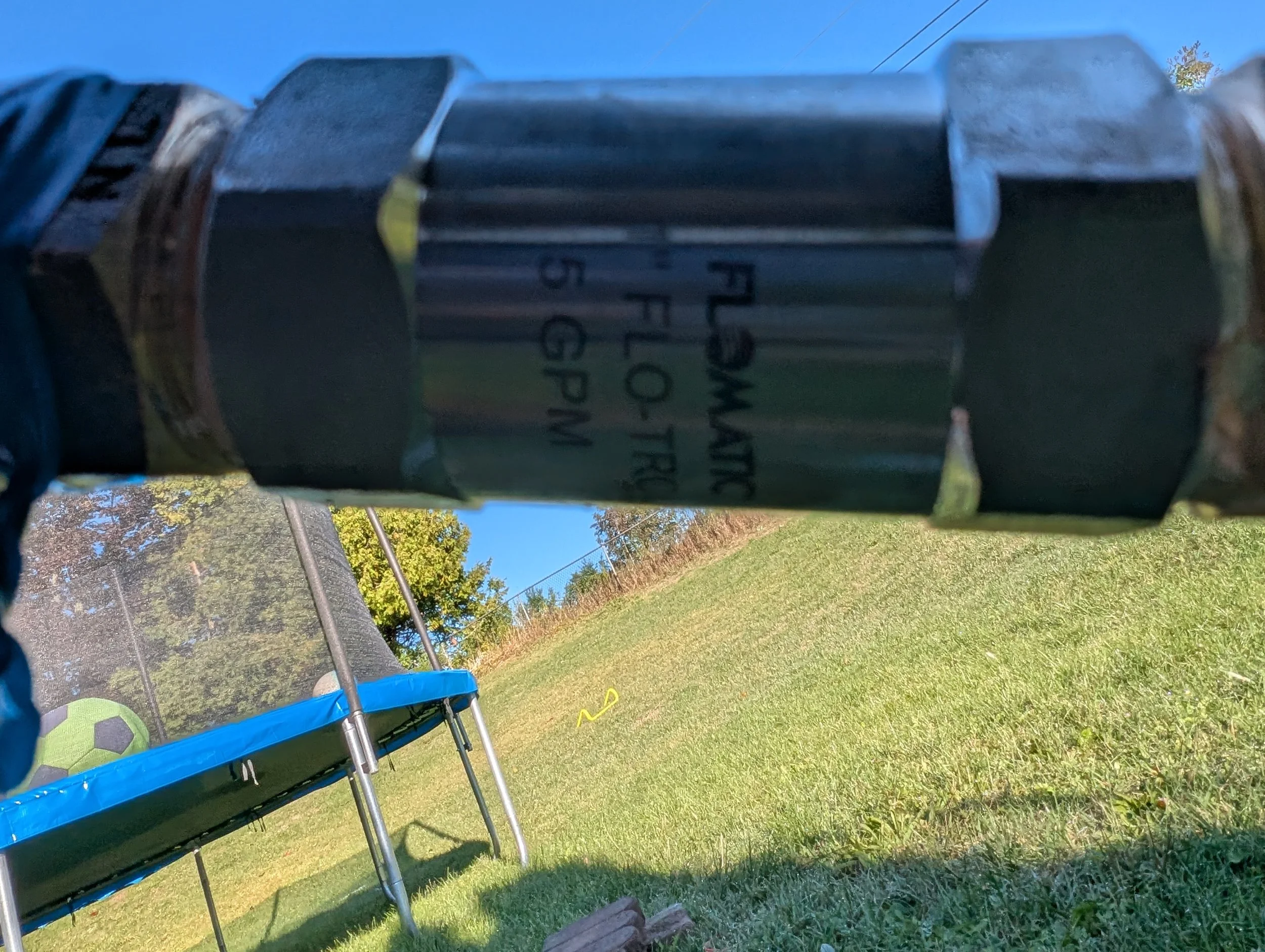 Close-up of a motorcycle handlebar with a 12-volt power socket, outdoors with grass and trees in the background.