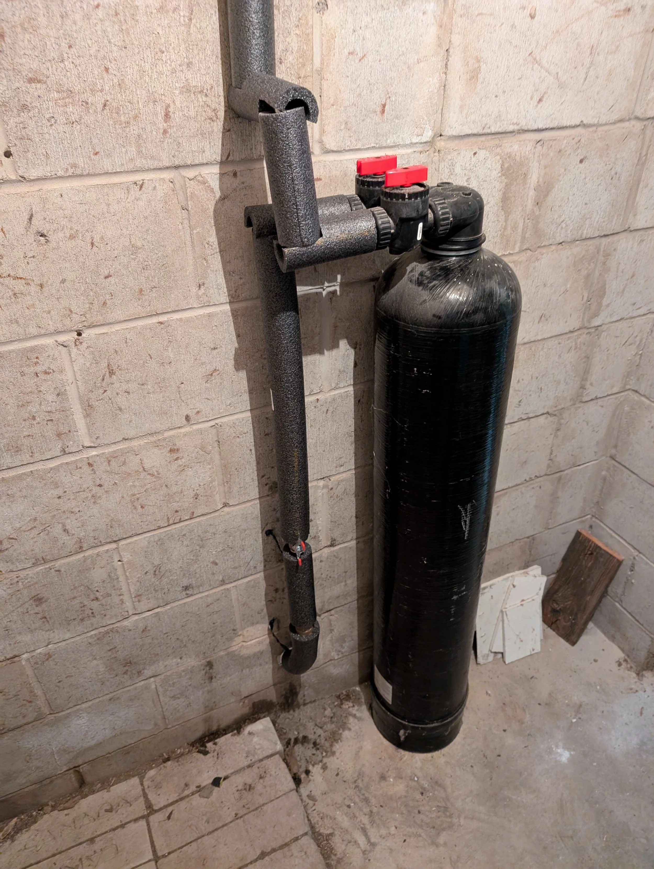 Close-up of a black water filtration or pressure tank with attached pipes and valves against a unfinished brick wall, in a basement or utility room.