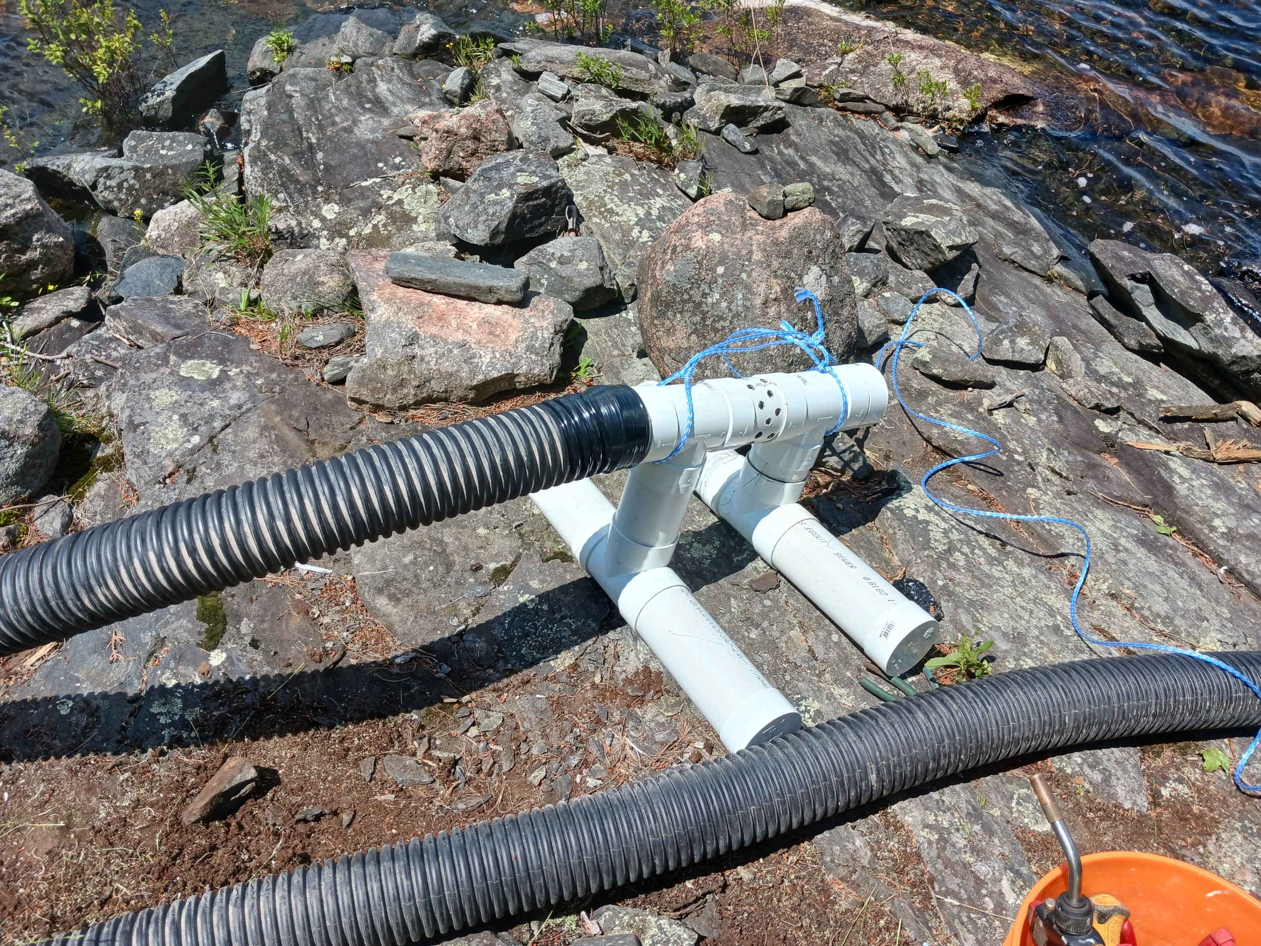 Leaking pipes, hoses, and equipment on rocky shoreline with water nearby.