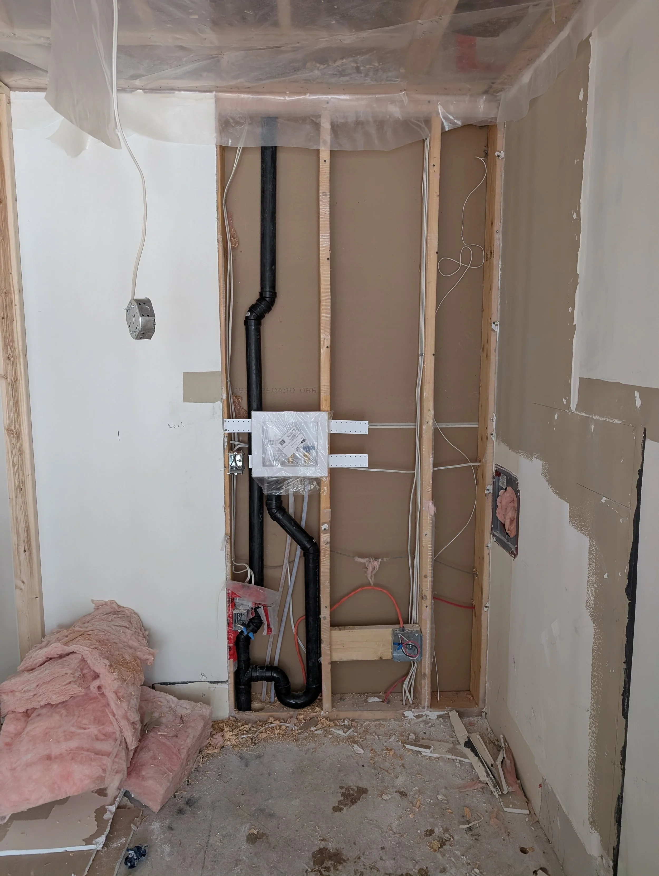 Construction room showing plumbing pipes, electrical wires, and unfinished walls with insulation and drywall.
