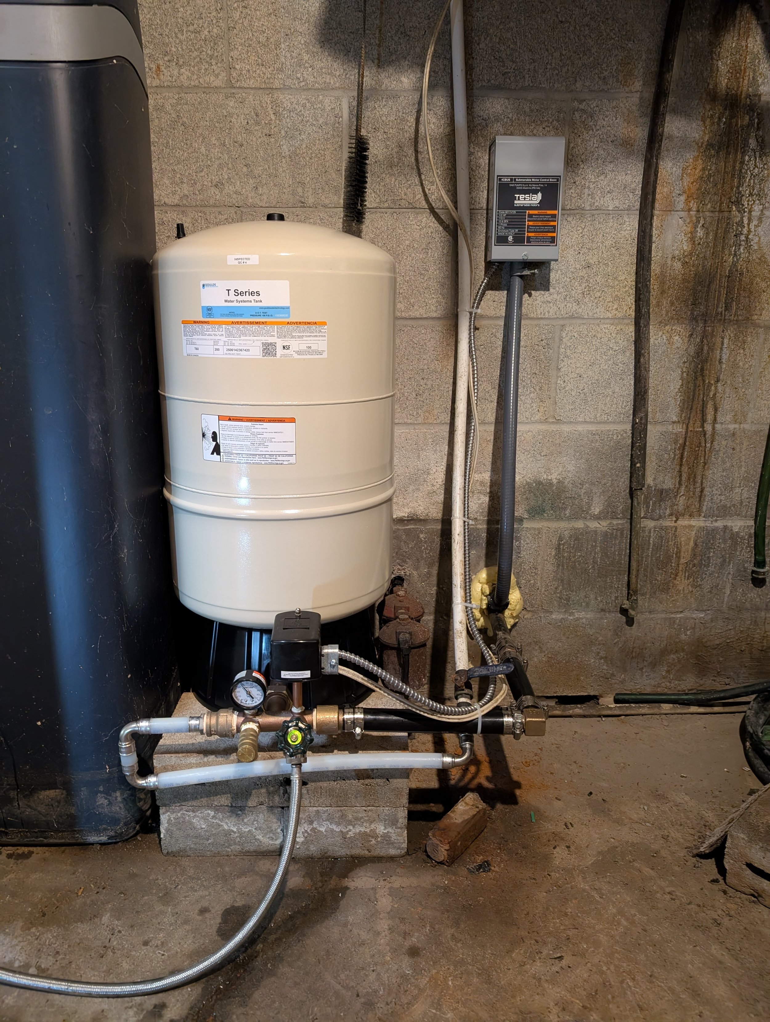 A water system setup with a white water tank, black tank, and various pipes and gauges along a concrete block wall.