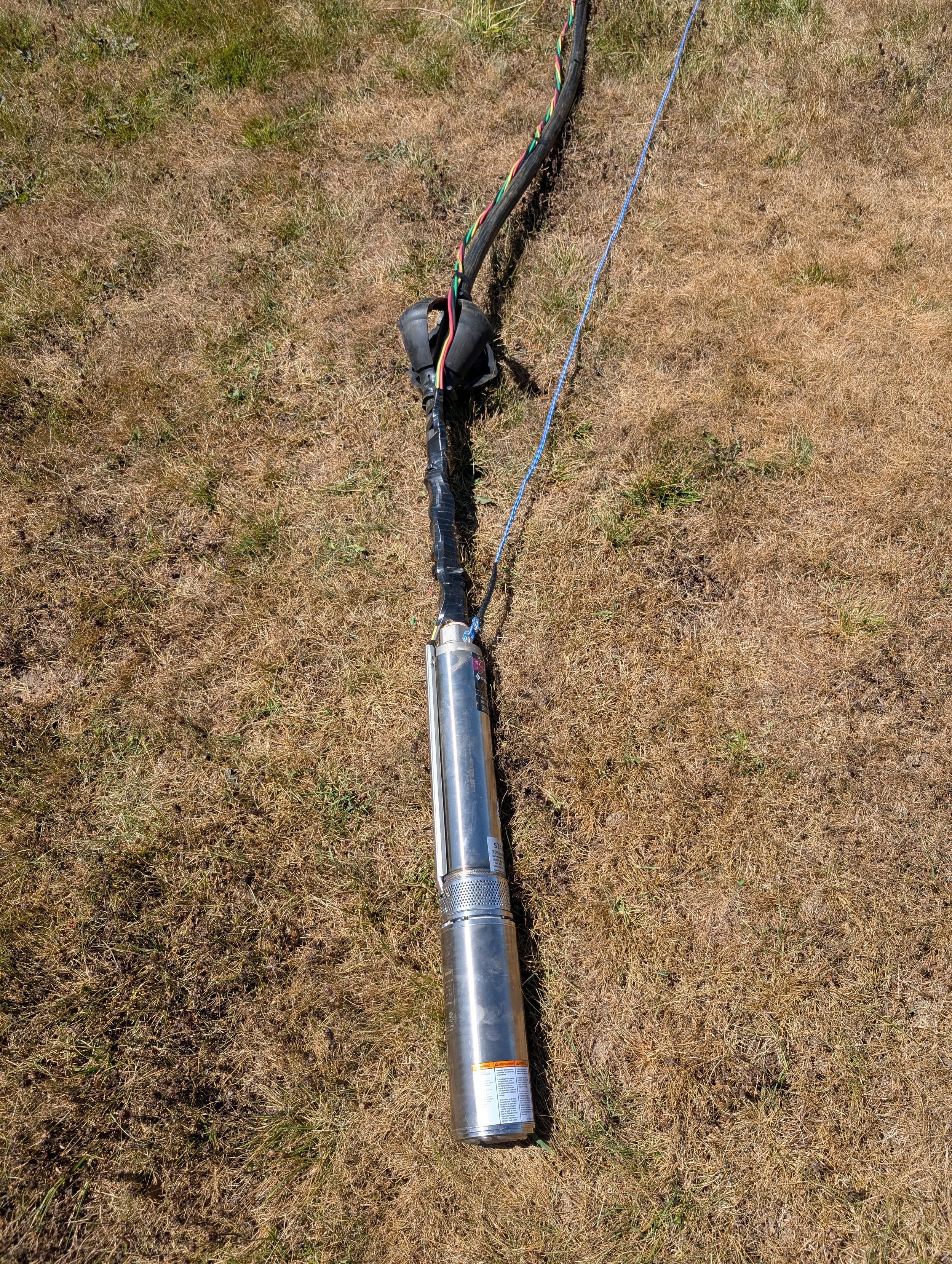 A large metal device, likely a piece of industrial or scientific equipment, lies on a grassy field. The device is long and cylindrical with wires and cables attached, and some labels and warnings are visible on its surface.
