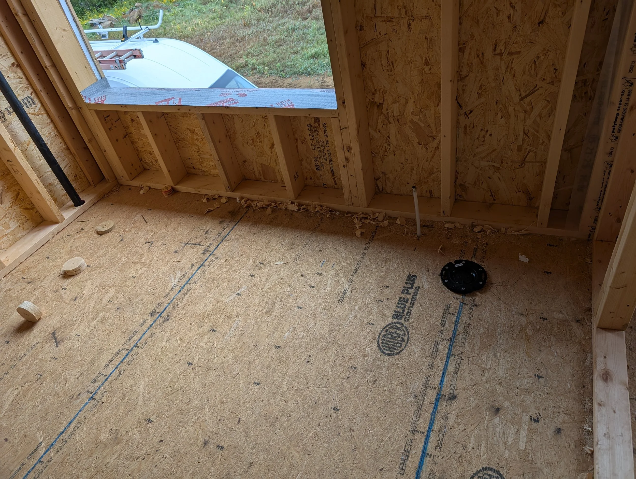 Interior view of a building under construction with exposed wooden framing, oriented strand board sheathing, and a small window showing a grassy outdoor area with a white vehicle outside.