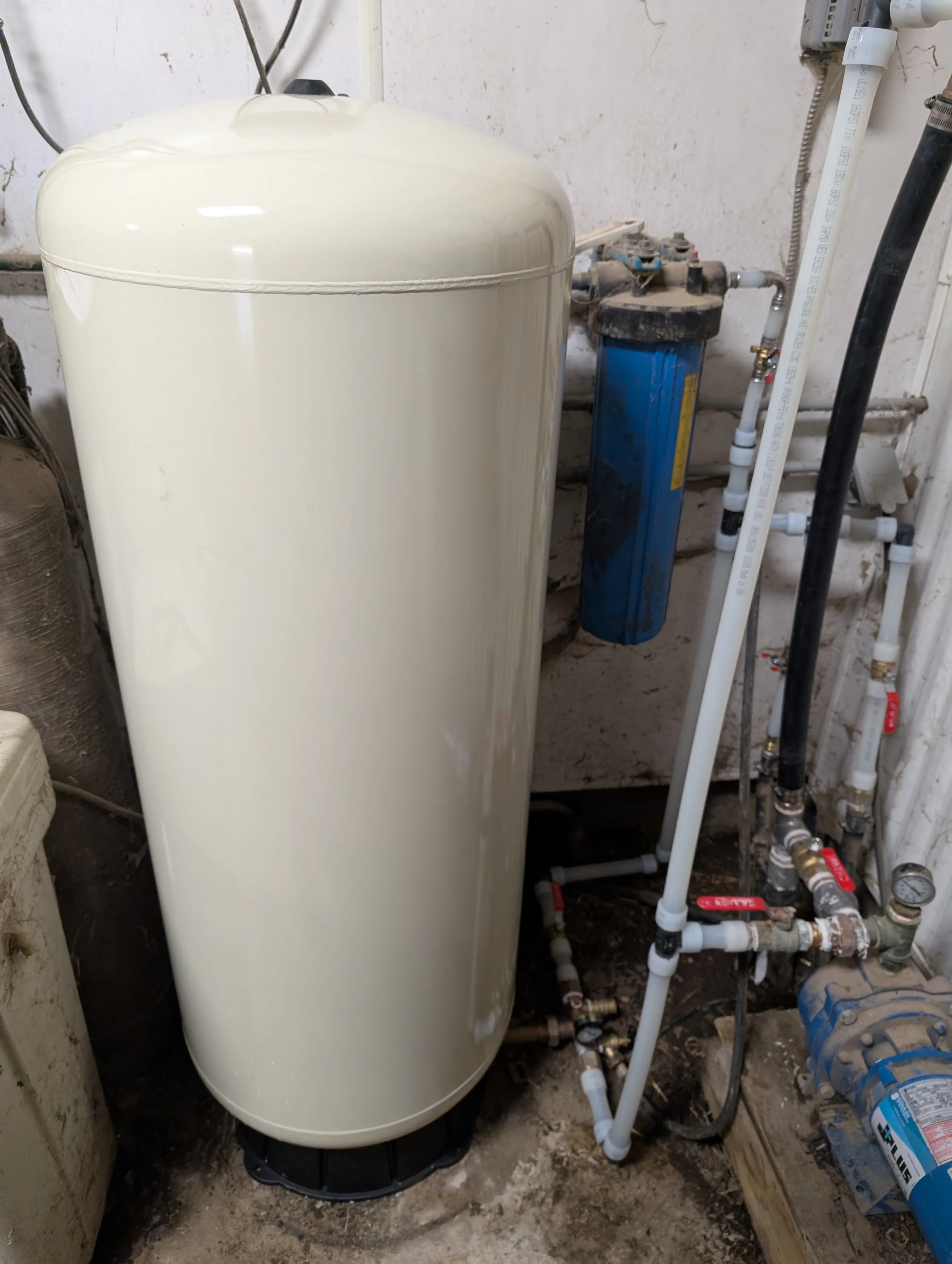 An image showing a water filtration system in a basement, including a large white water softener tank, a blue filter, and various pipes and gauges.