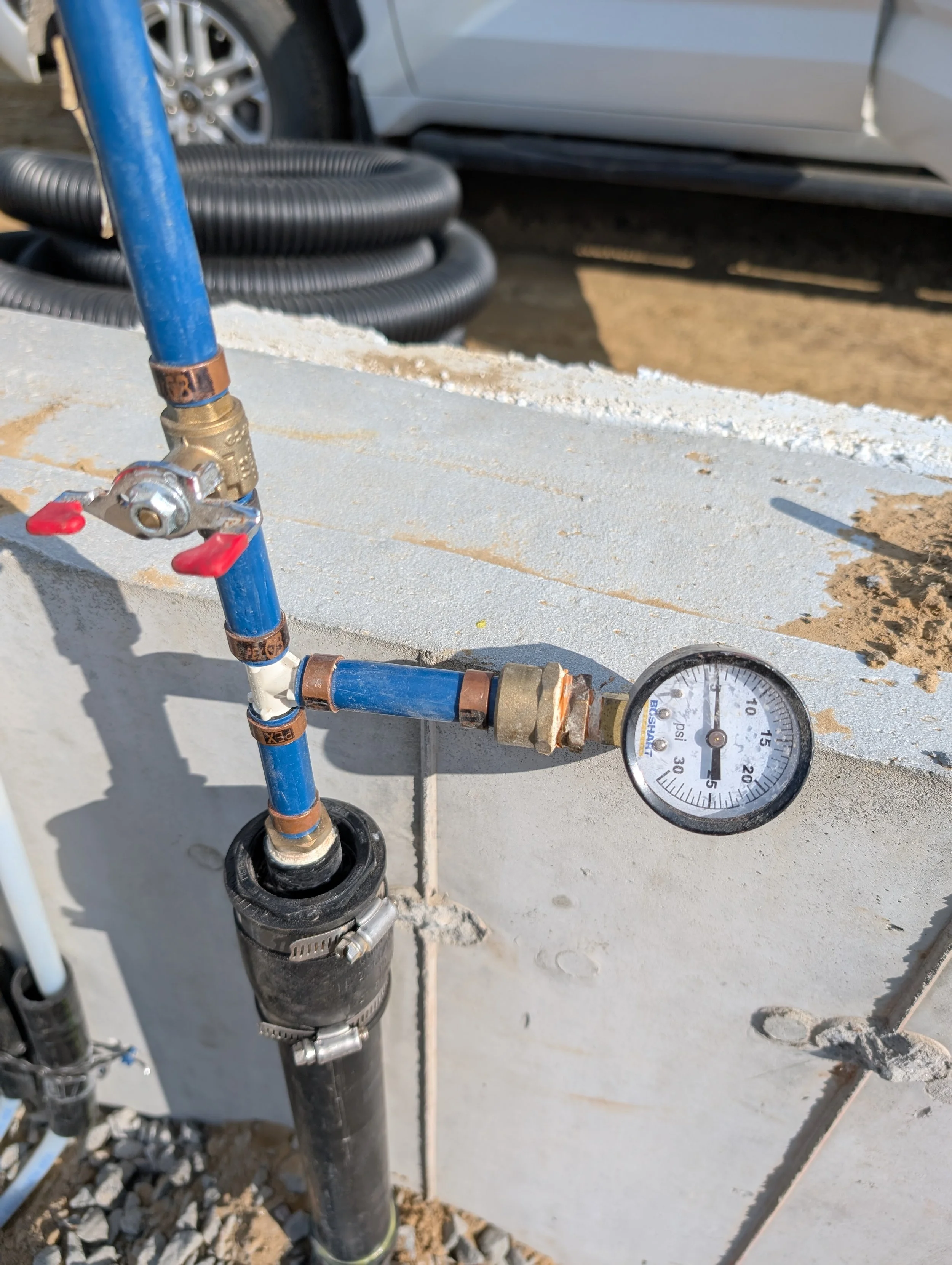Close-up of a water pressure gauge connected to black and blue piping on a construction site, with a coiled black hose and a white vehicle in the background.