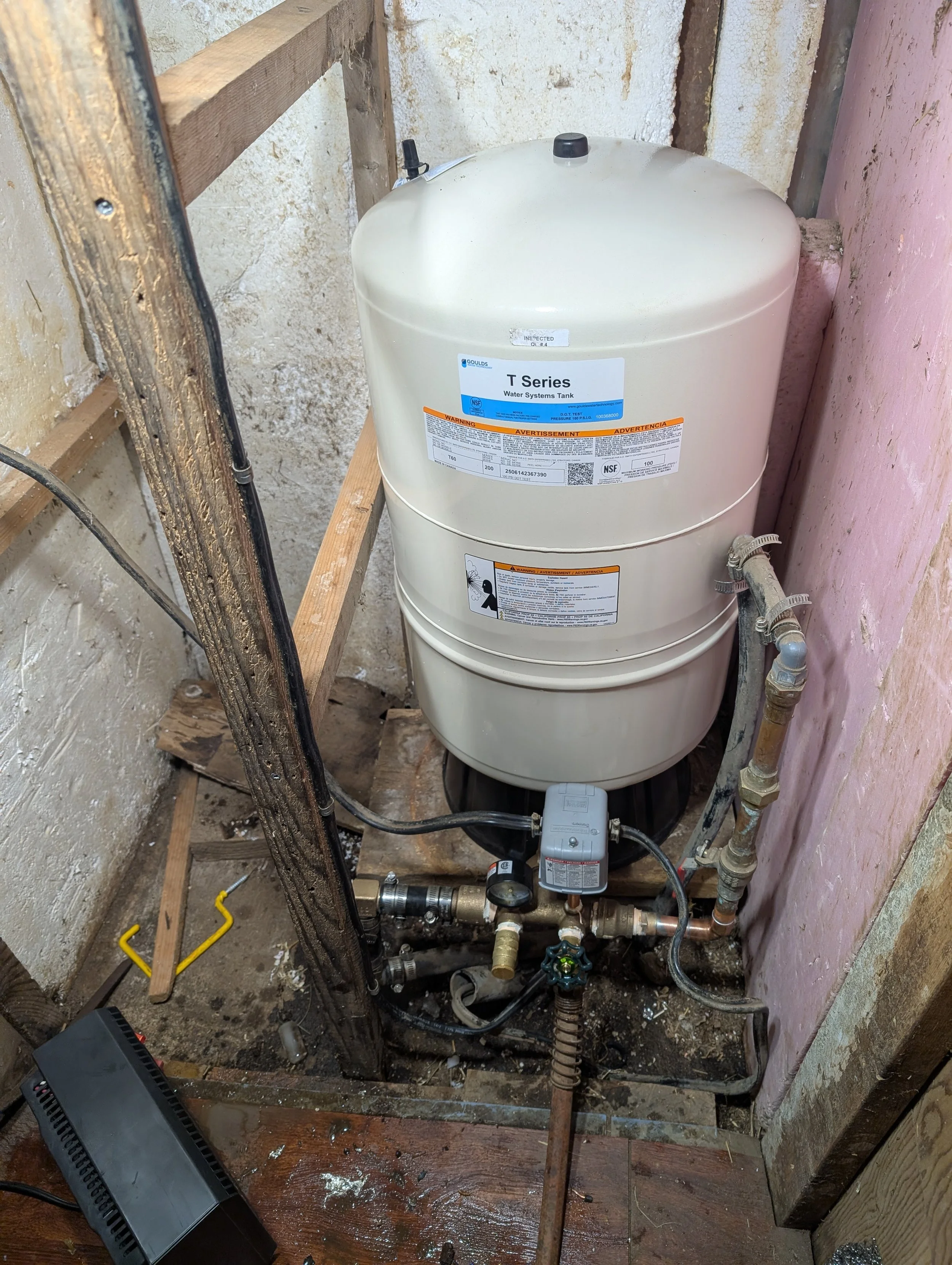 Water system tank labeled 'T Series' installed in a construction area with exposed pipes, a pressure valve, and a wooden frame surrounding it.