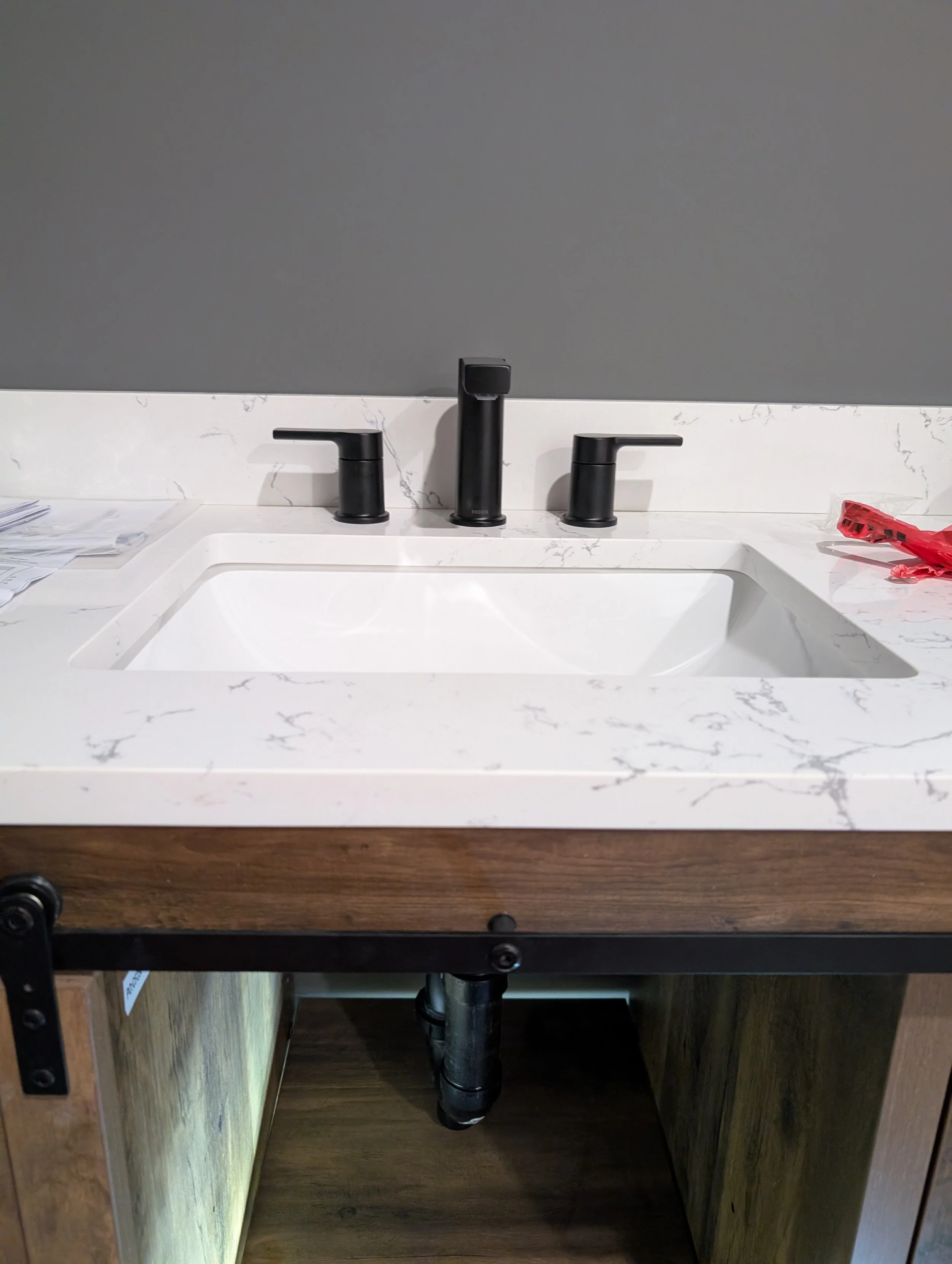 A bathroom vanity with a white marble countertop, black faucet, and black handles. There are some papers on the left side and a red object on the right side. The vanity cabinet is made of dark wood.