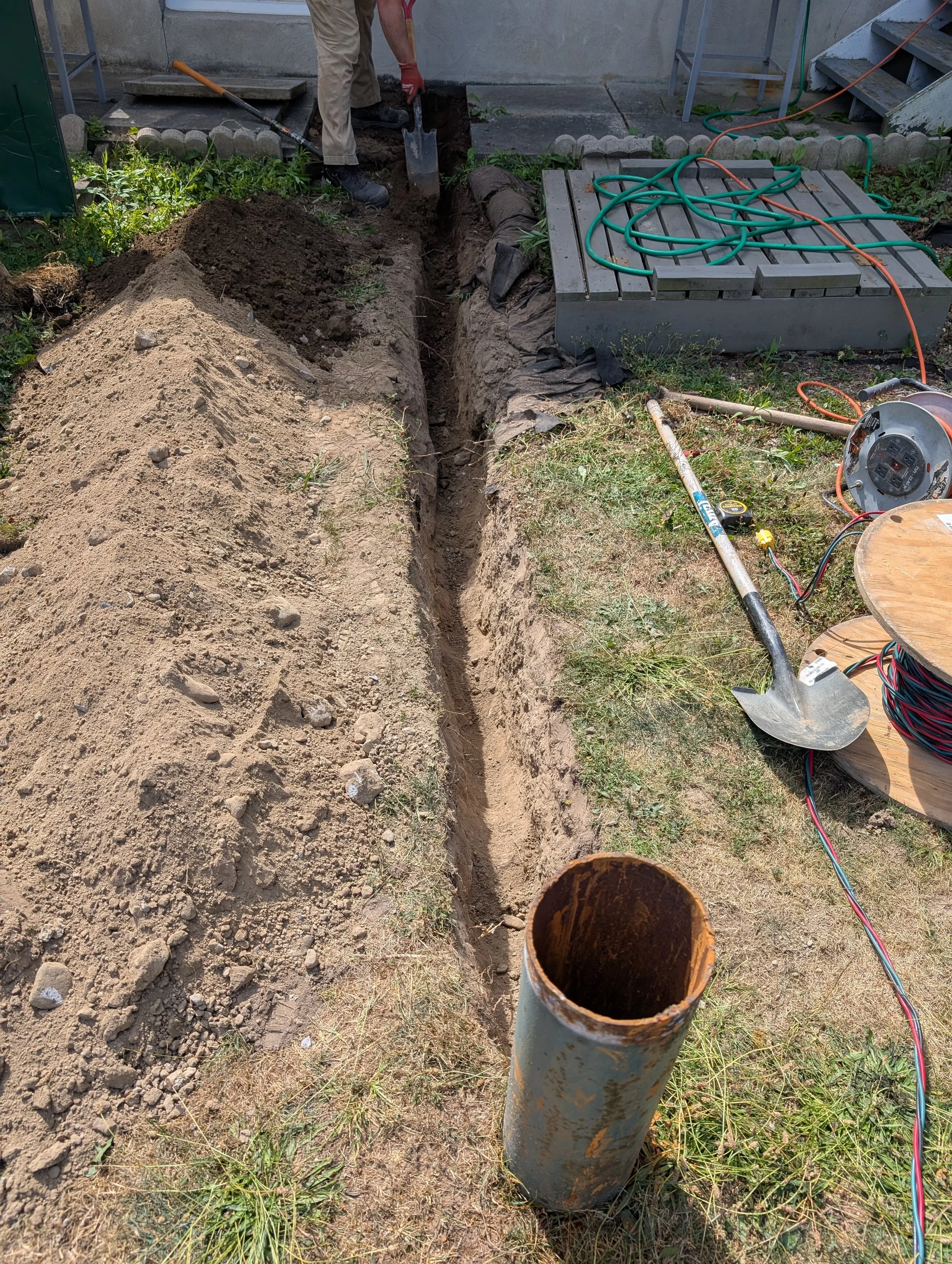 A trench dug in the ground for underground work, with a worker shoveling dirt nearby, and various tools and cables on the grass and a gray container with hoses on it.