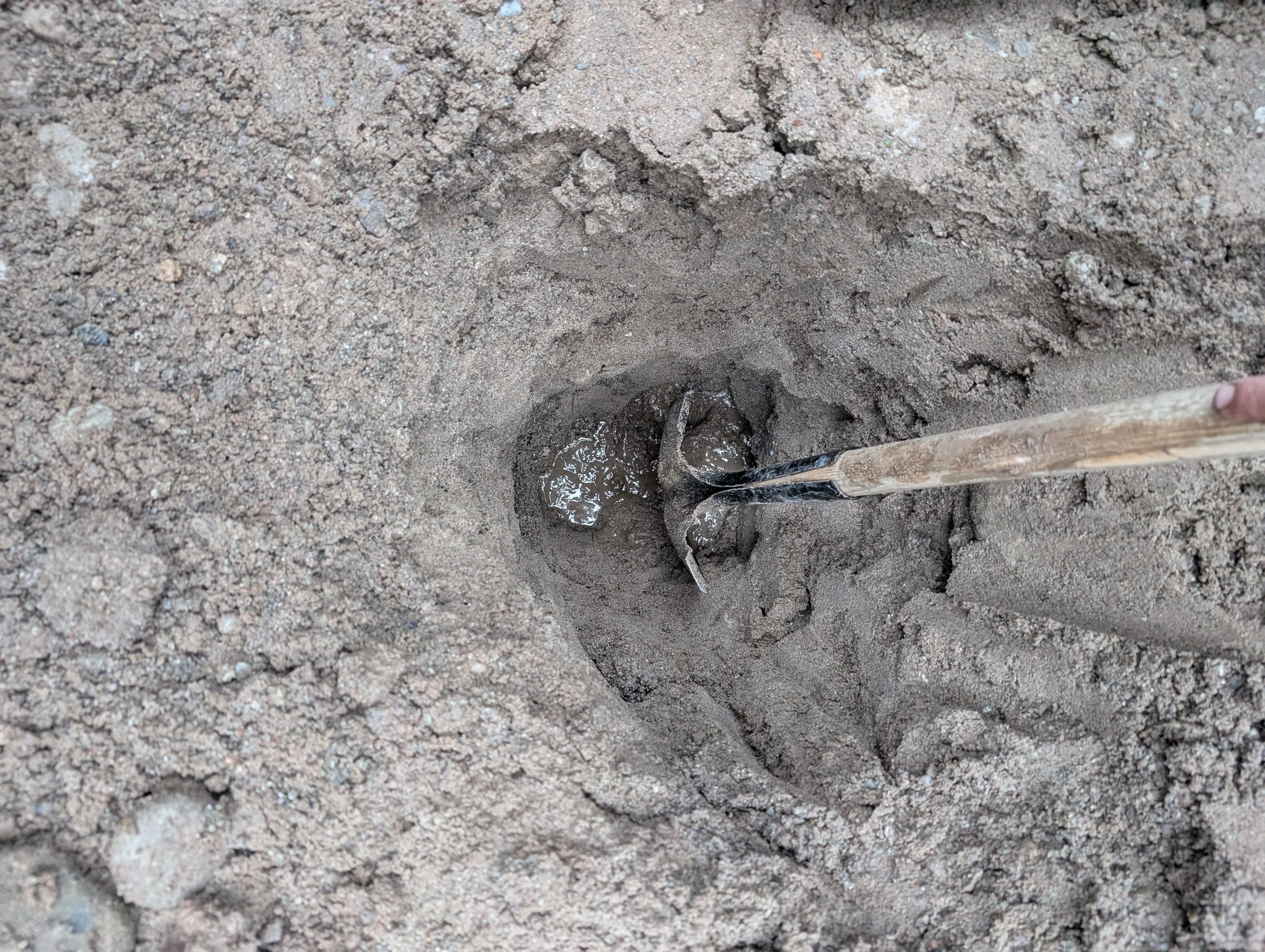 A small hole in the ground with water and a hand tool, possibly a shovel, next to it on a dirt surface.