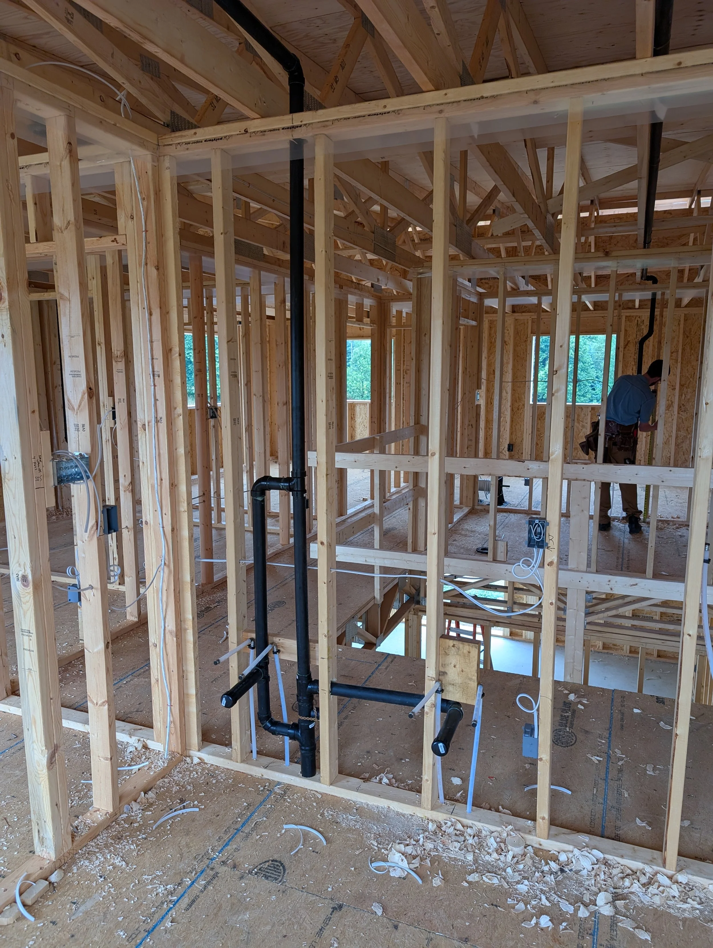 The image shows a residential construction site with wooden framing, exposed pipes, electrical wiring, and a worker inspecting the work. The interior walls are not yet finished.