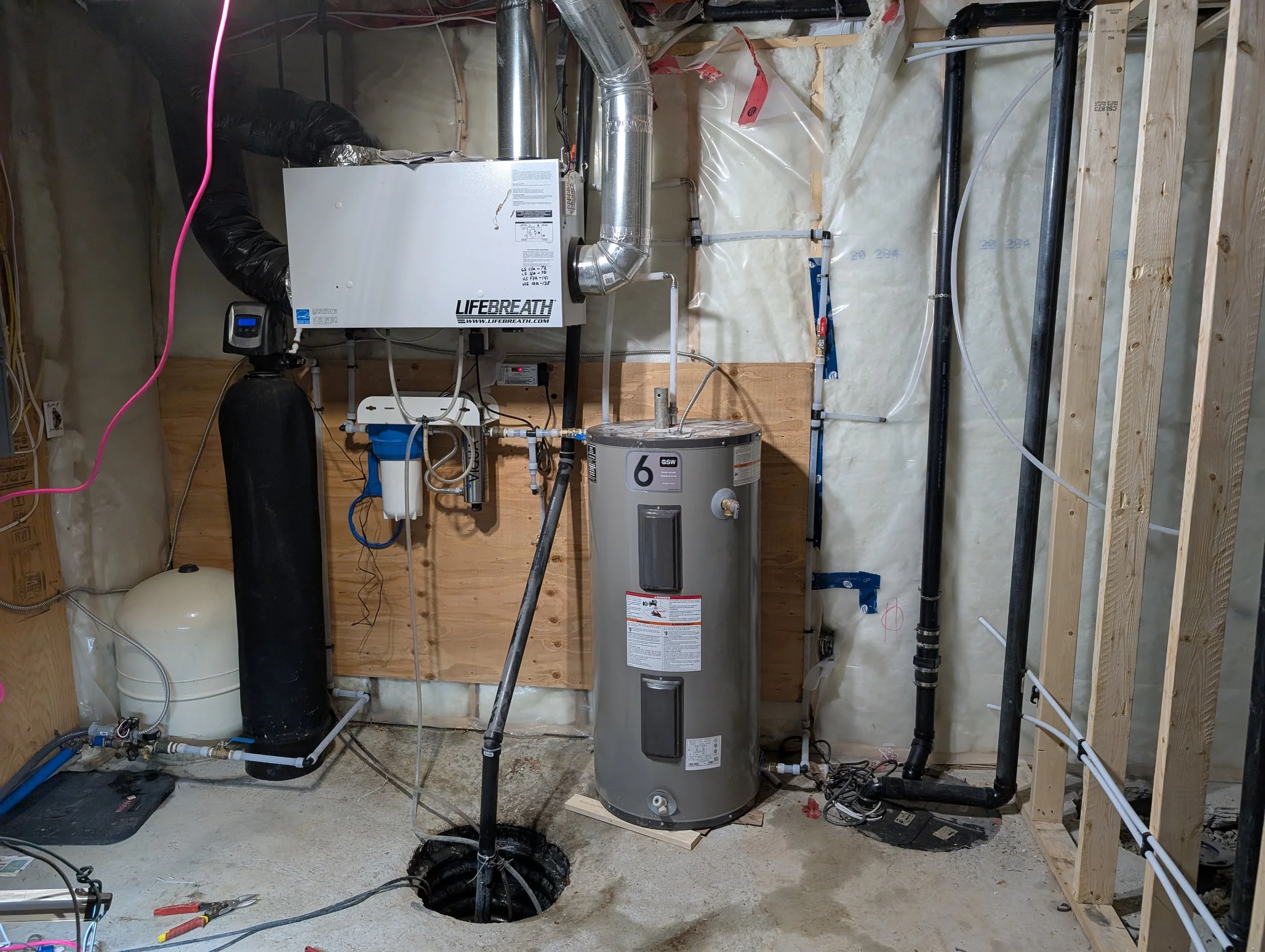 An unfinished basement with exposed wooden framing, insulation, and plumbing pipes. A water heater and HVAC system are visible, with electrical wires and tools lying on the floor.