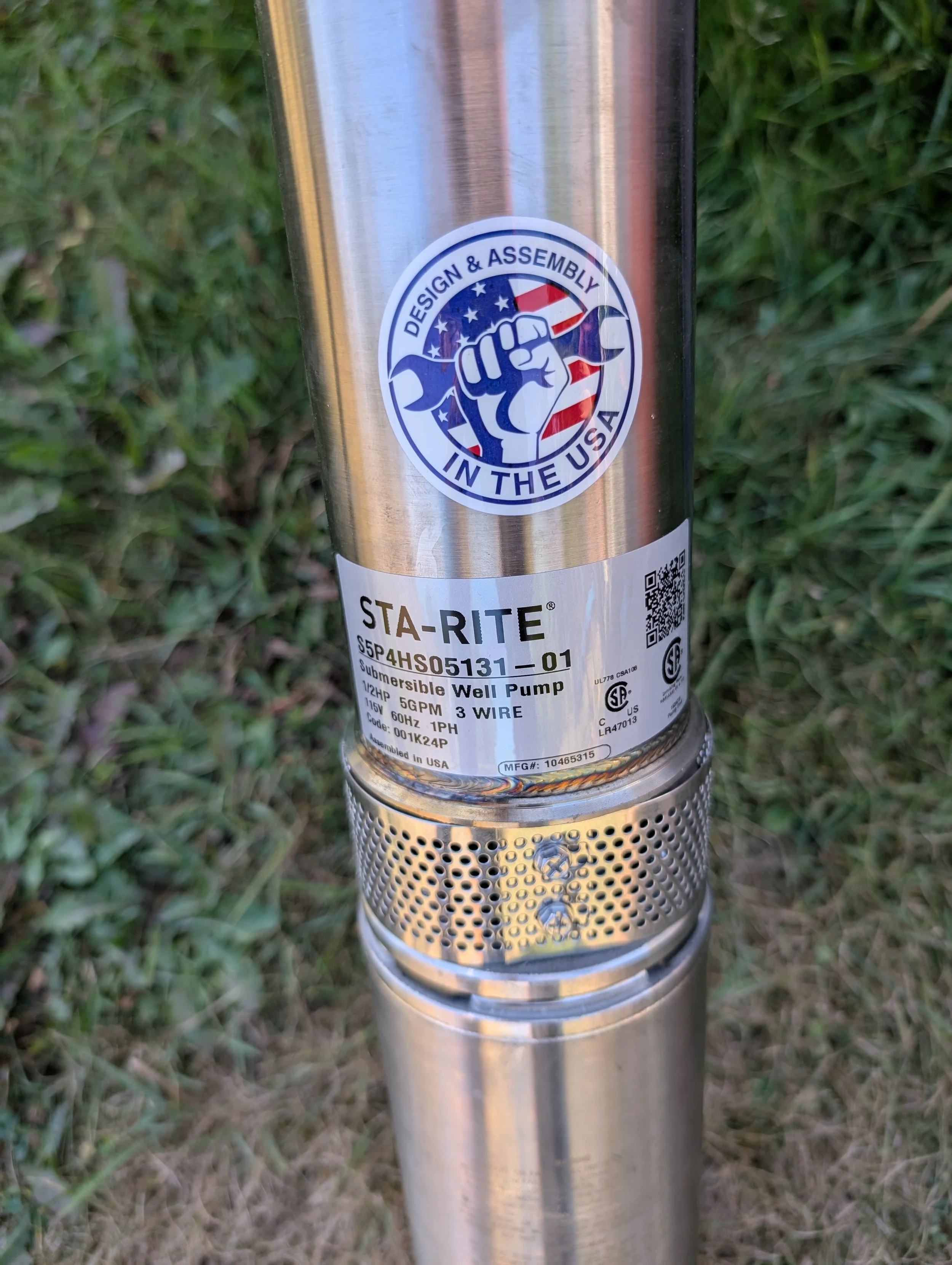Close-up of a stainless steel submersible well pump with a circular sticker that reads 'Design & Assembly in the USA' and features a raised fist holding a wrench, set against a grassy background.