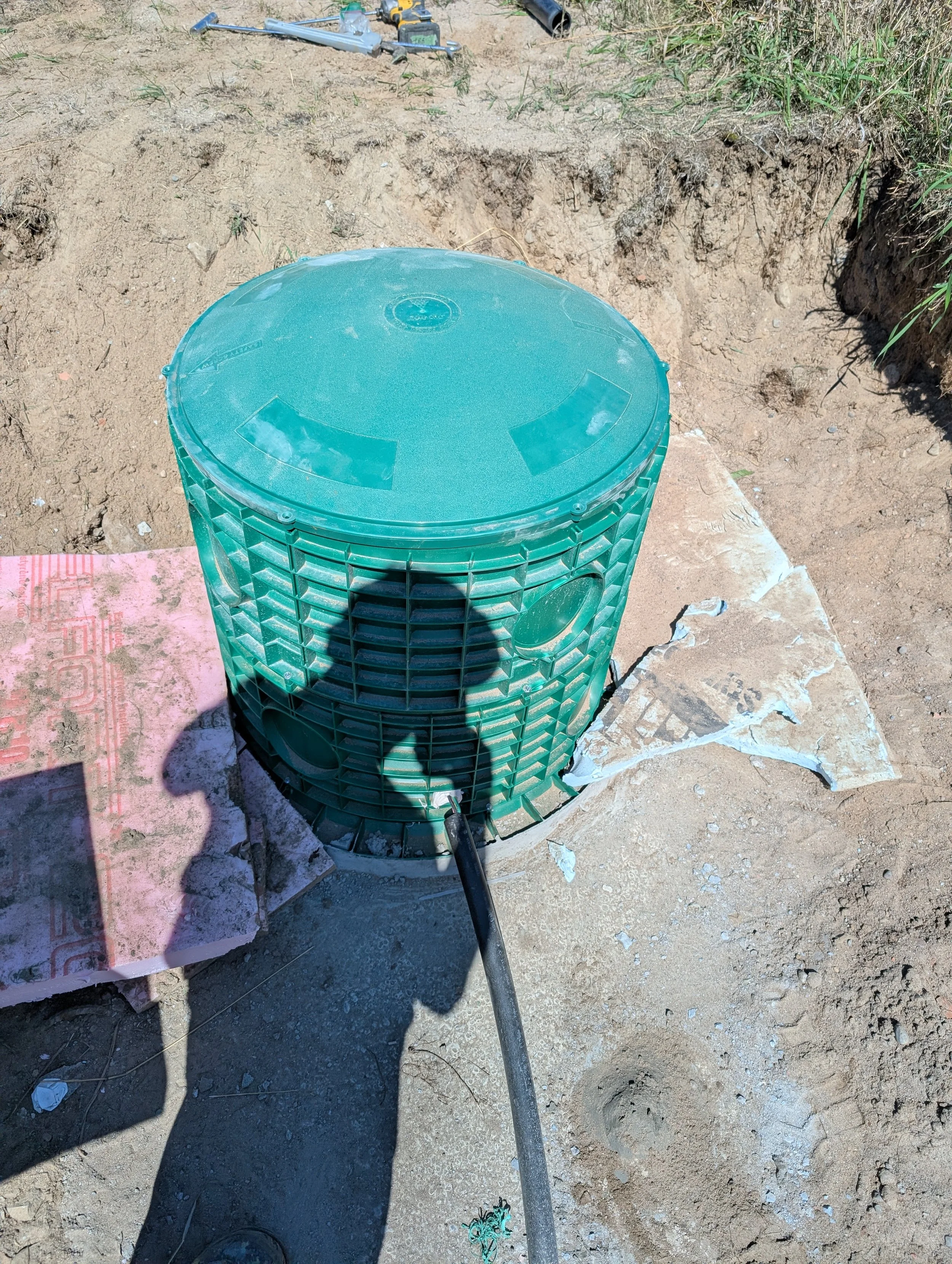 A green bucket with a lid is placed on the ground outdoors on a construction site with dirt and rocks. A black cable runs from the bucket into a hole in the ground. In the background, there are scattered tools, including a drill and other equipment, 
