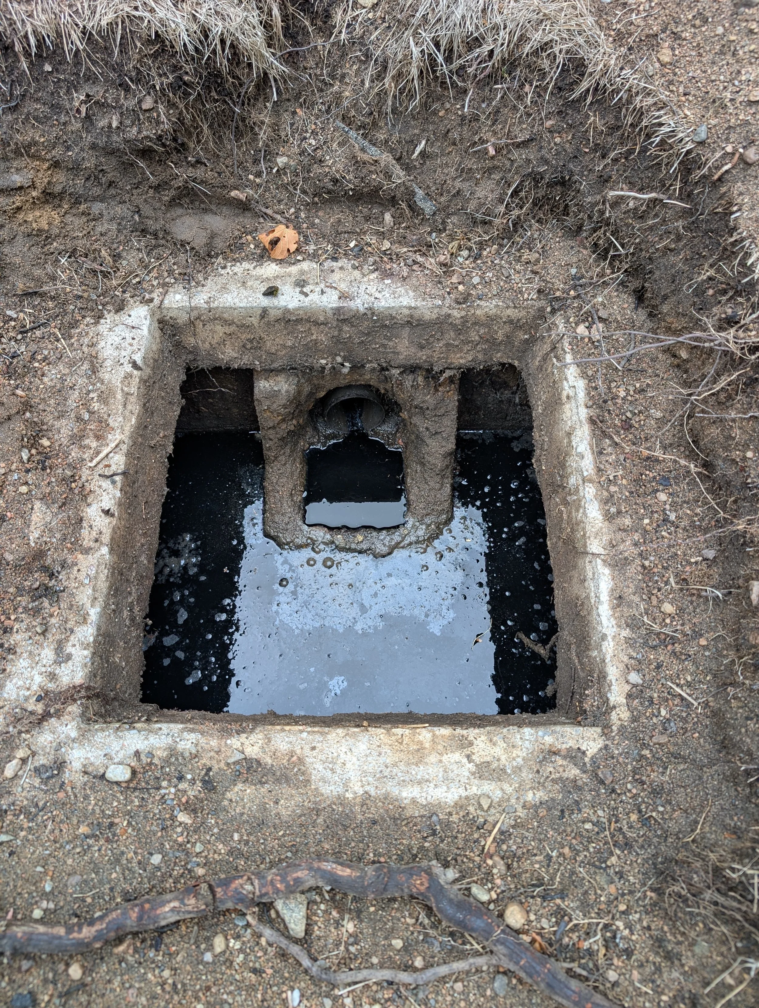 An outdoor concrete drain with water flowing through it, embedded in rocky dirt ground with some sticks and a dead leaf on the surface.