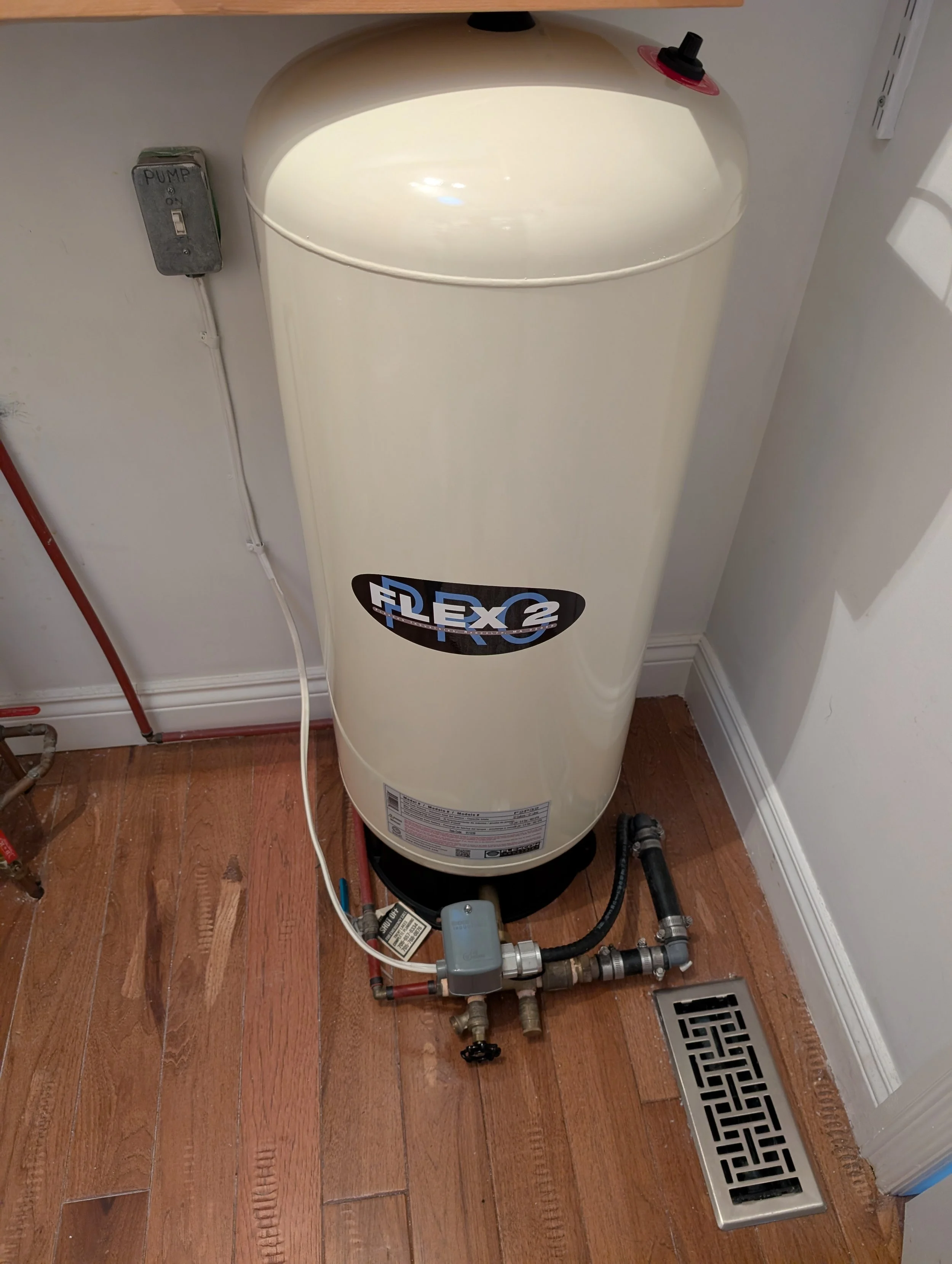 A water heater installed in a home with connecting pipes, a drain grate, and a nearby control box on the wall.