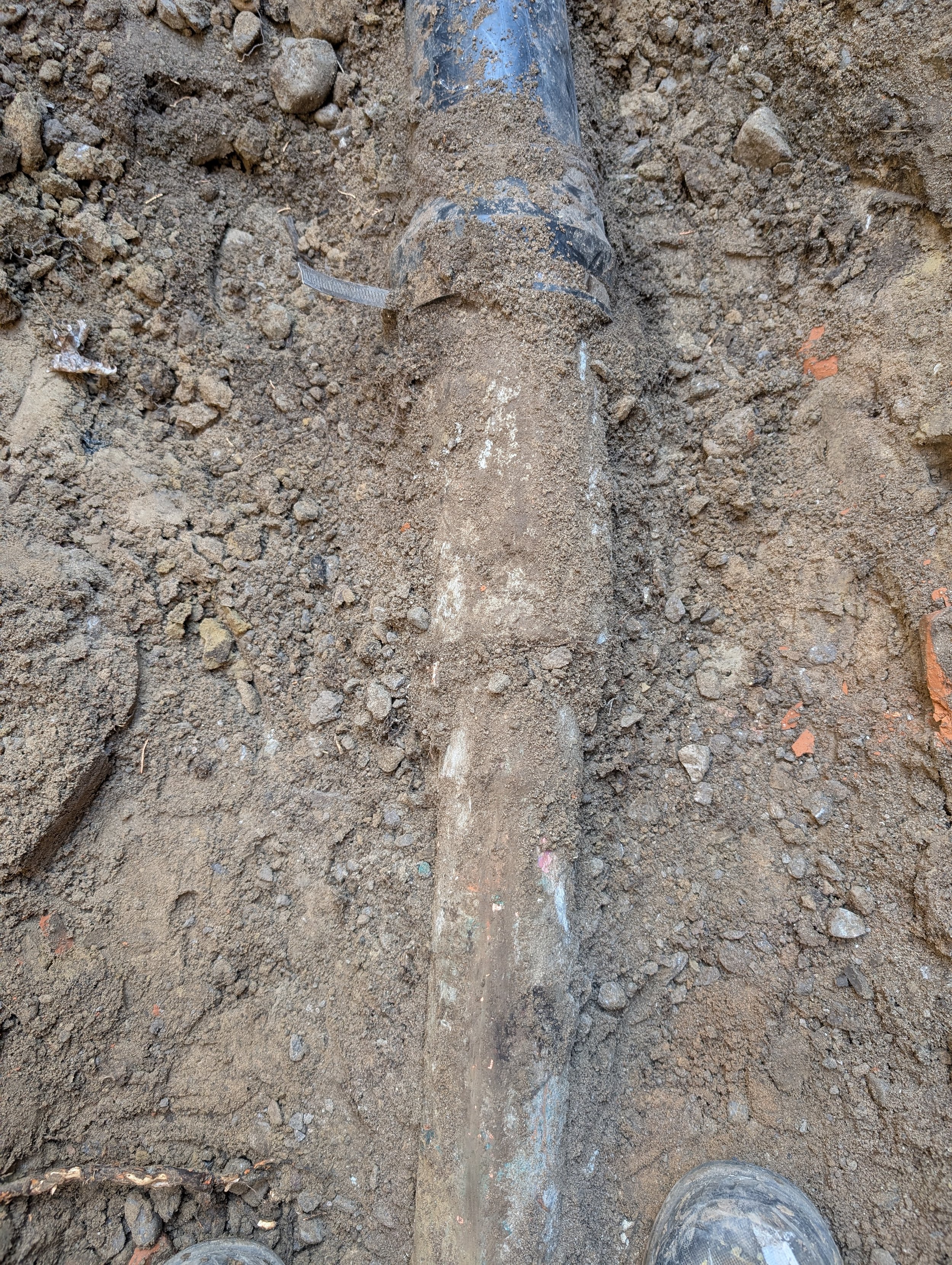 A dirt ground with a fallen black pipe or hose partially buried in the soil and gravel. The perspective is from someone wearing muddy work boots looking down at the pipe.