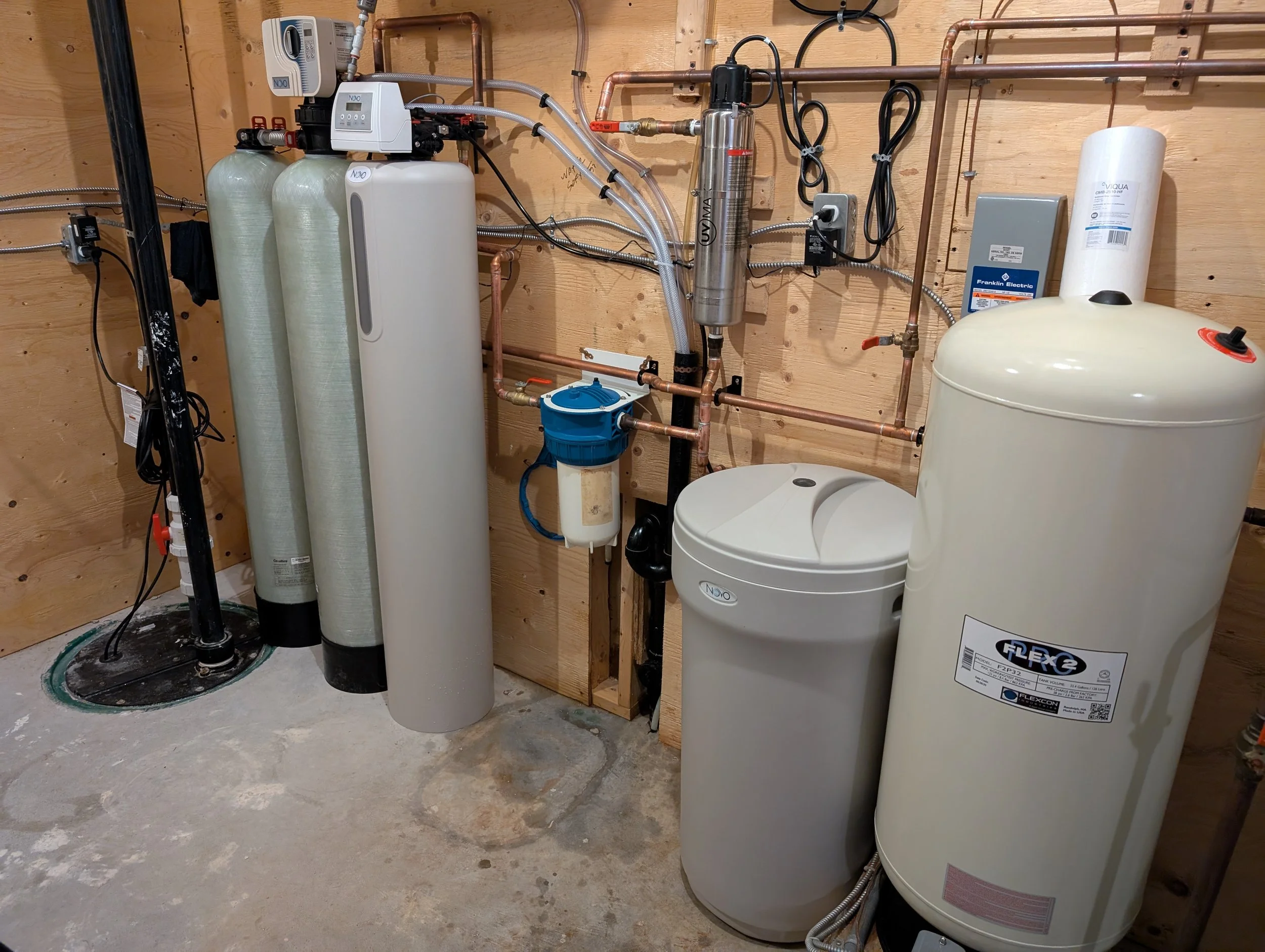 A basement or utility room with water filtration and pressure systems, including large water softener tanks, a tank for reverse osmosis, and various copper plumbing pipes.