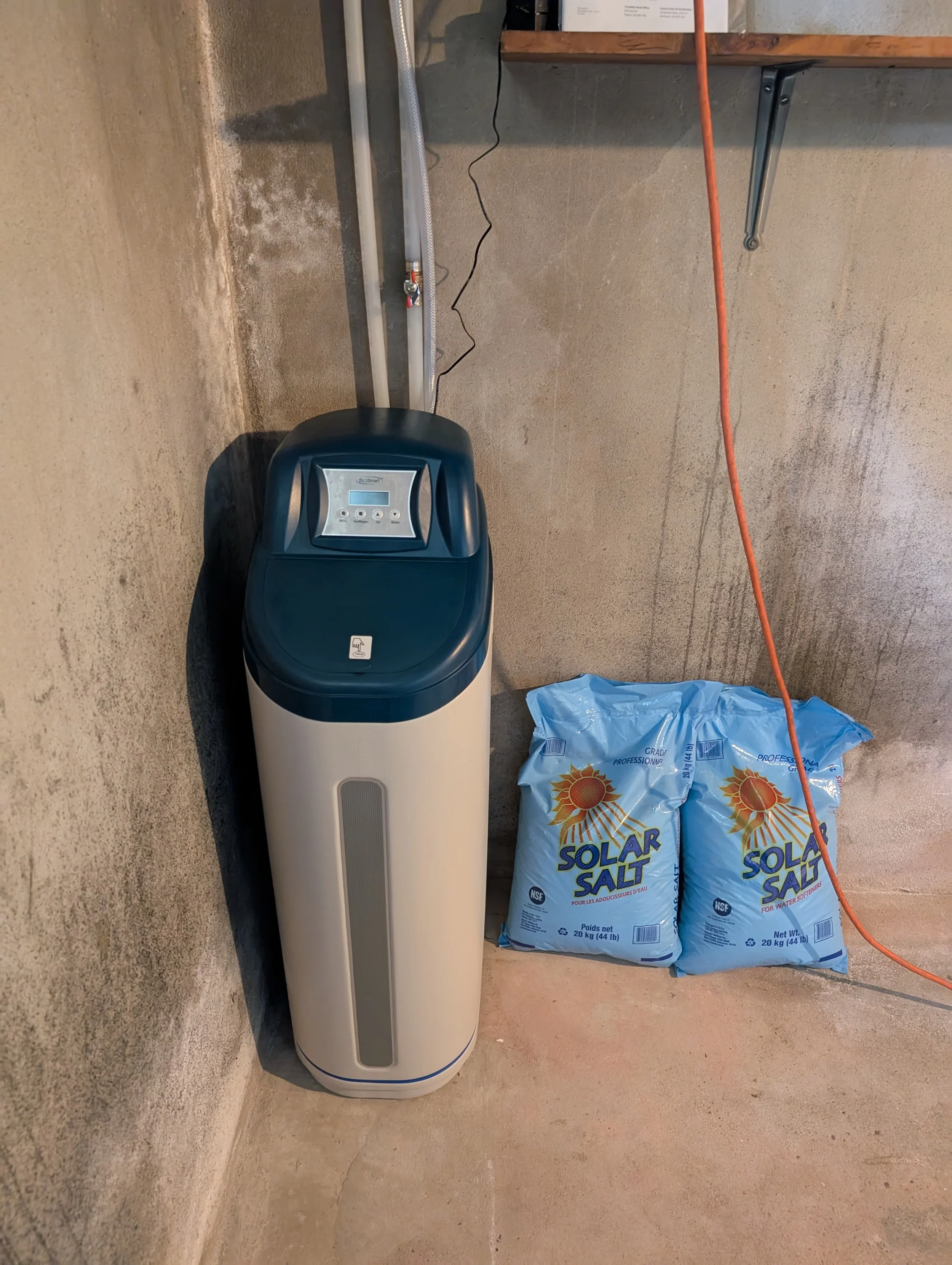 A water softener machine with a display screen is positioned against a concrete wall in a basement or utility room. To the right, there are two large bags labeled 'Solar Salt,' which are used for water softening. An orange extension cord runs across 