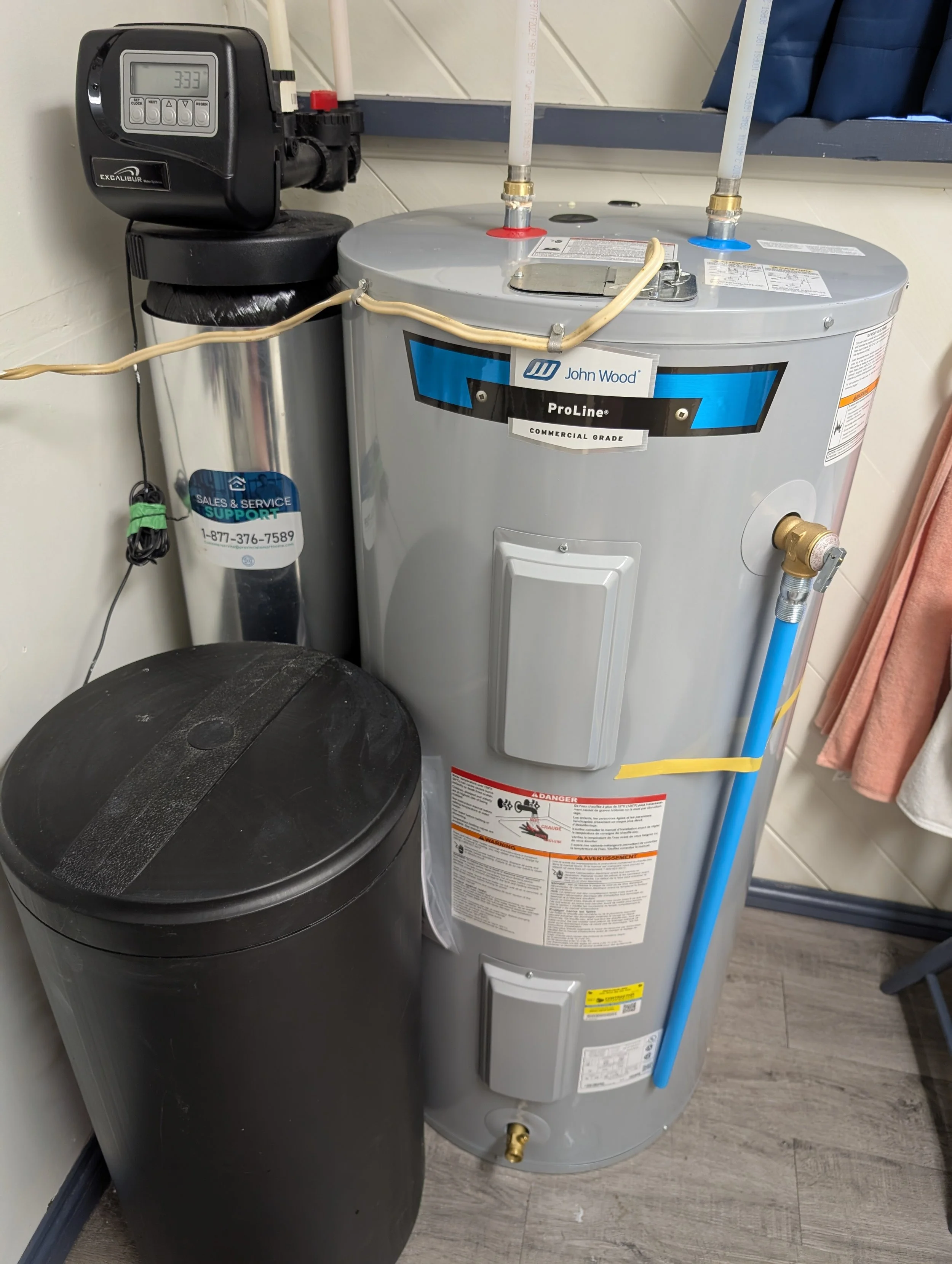 A water heater with pipes and a digital control panel, positioned next to a black cylinder with a label, in a room with wood flooring and a rod with a pink cloth hanging on the right.