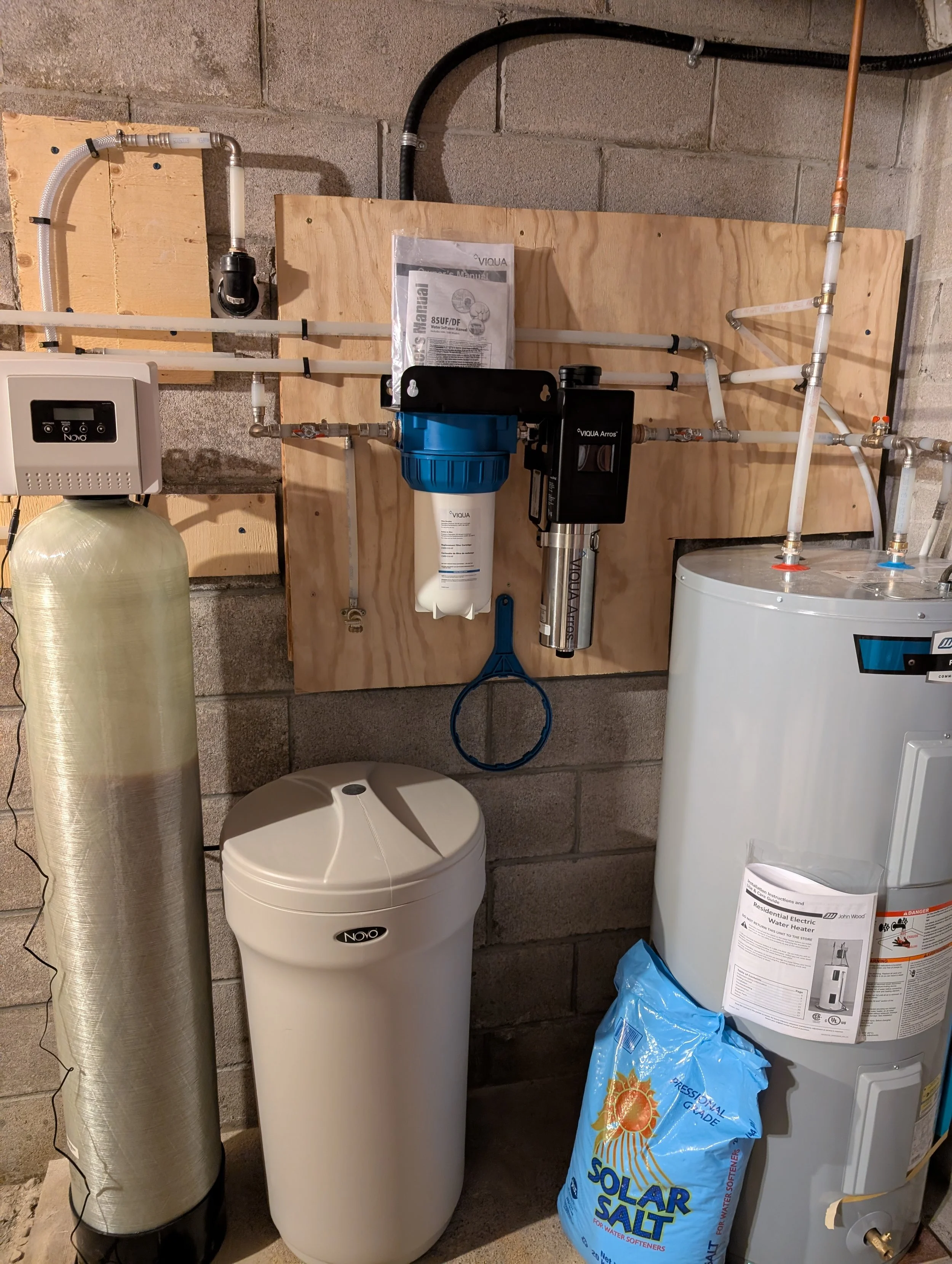 A water softener system with a salt storage container, a reverse osmosis water filter, and plumbing pipes against a concrete wall in a utility room.