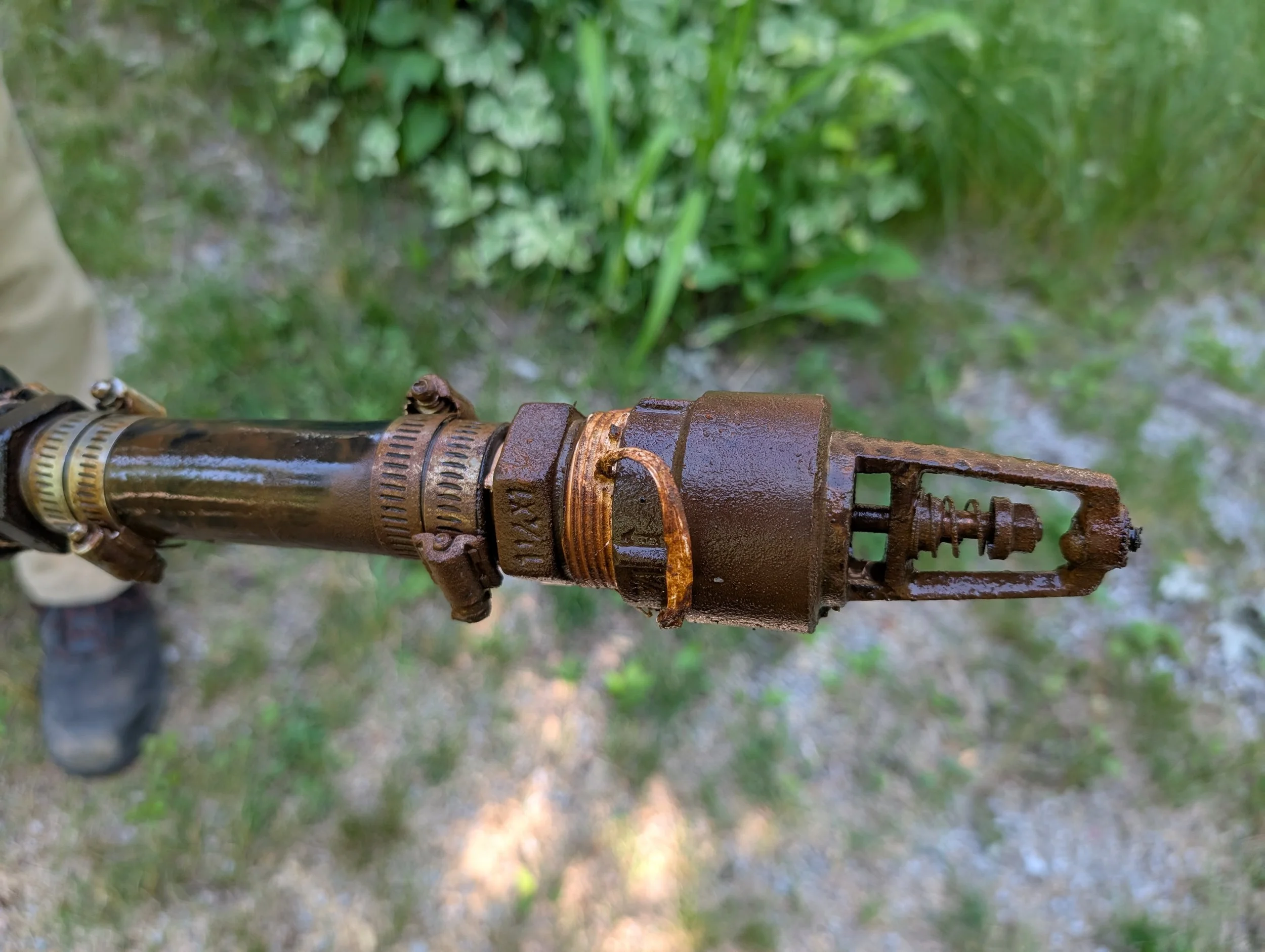 Close-up of rusted mechanical pipe or valve with green blurred foliage in the background.