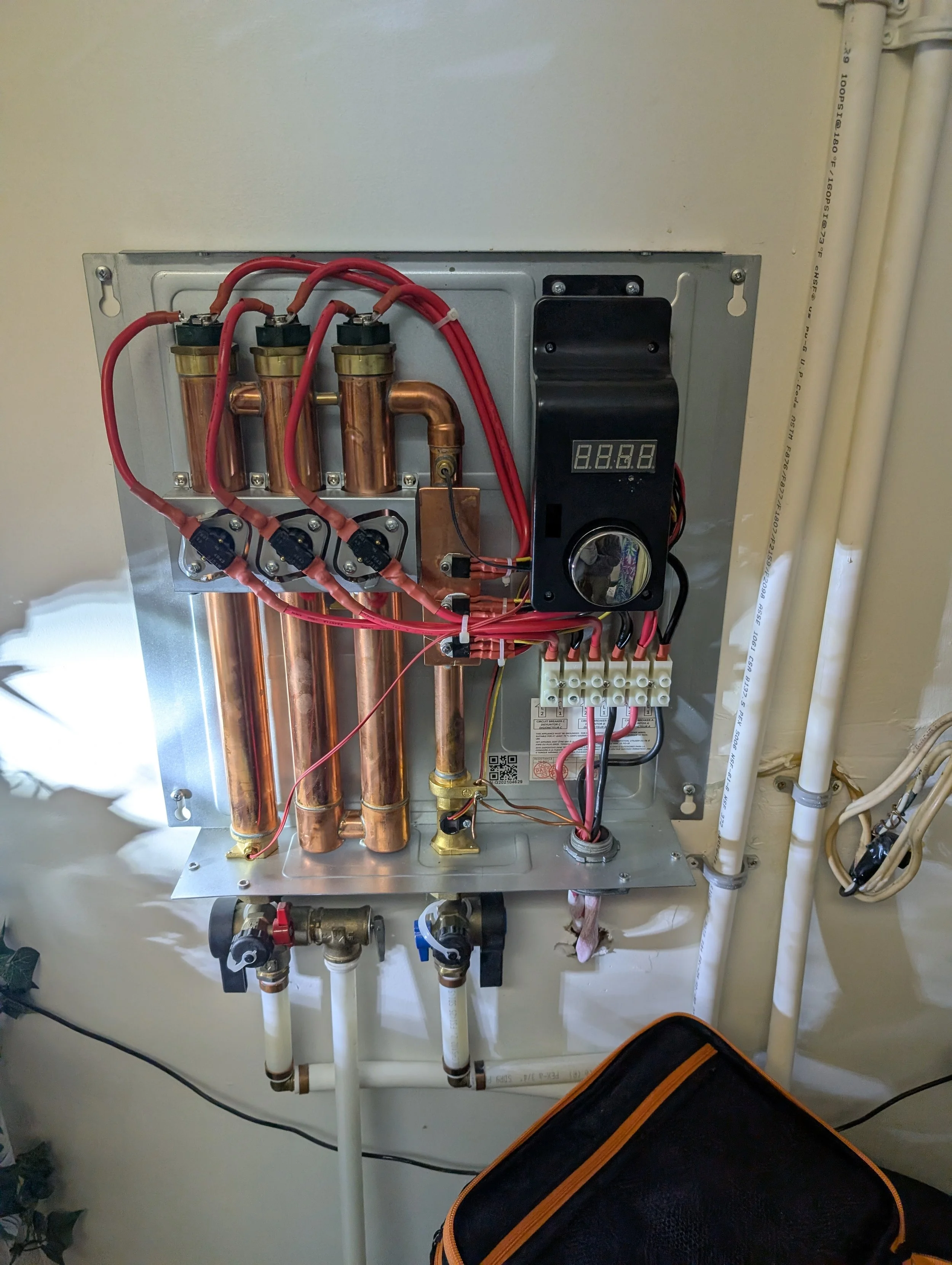 An electrical or plumbing control panel with copper pipes, wiring, a digital display, and valves, mounted on a wall with nearby pipes.