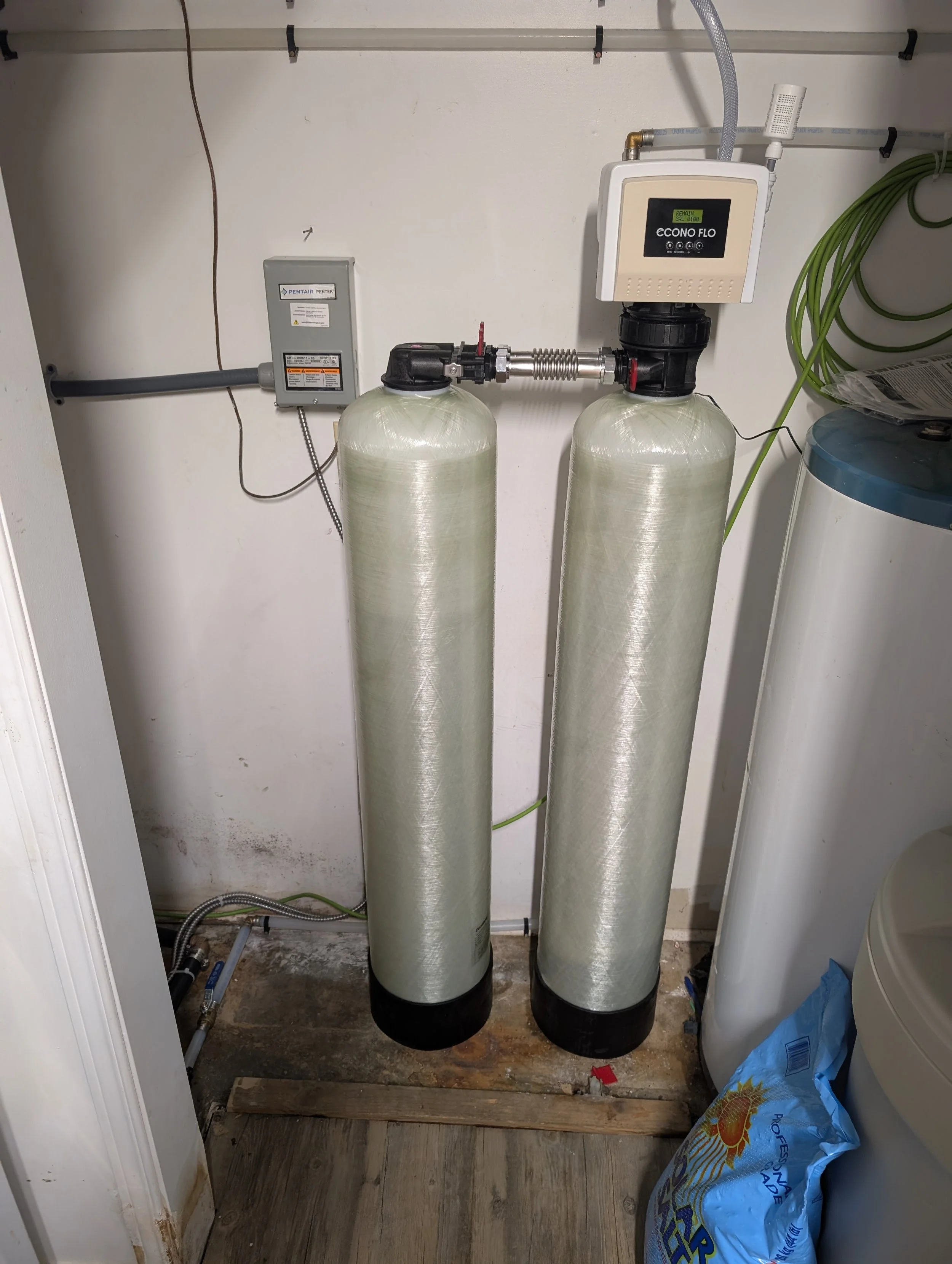 Two large water softener tanks are installed in a laundry room. The tanks are connected with pipes and a control head on top of the right tank. To the right side, there is a blue bag of water softener salt, and to the left, a small gray electrical bo