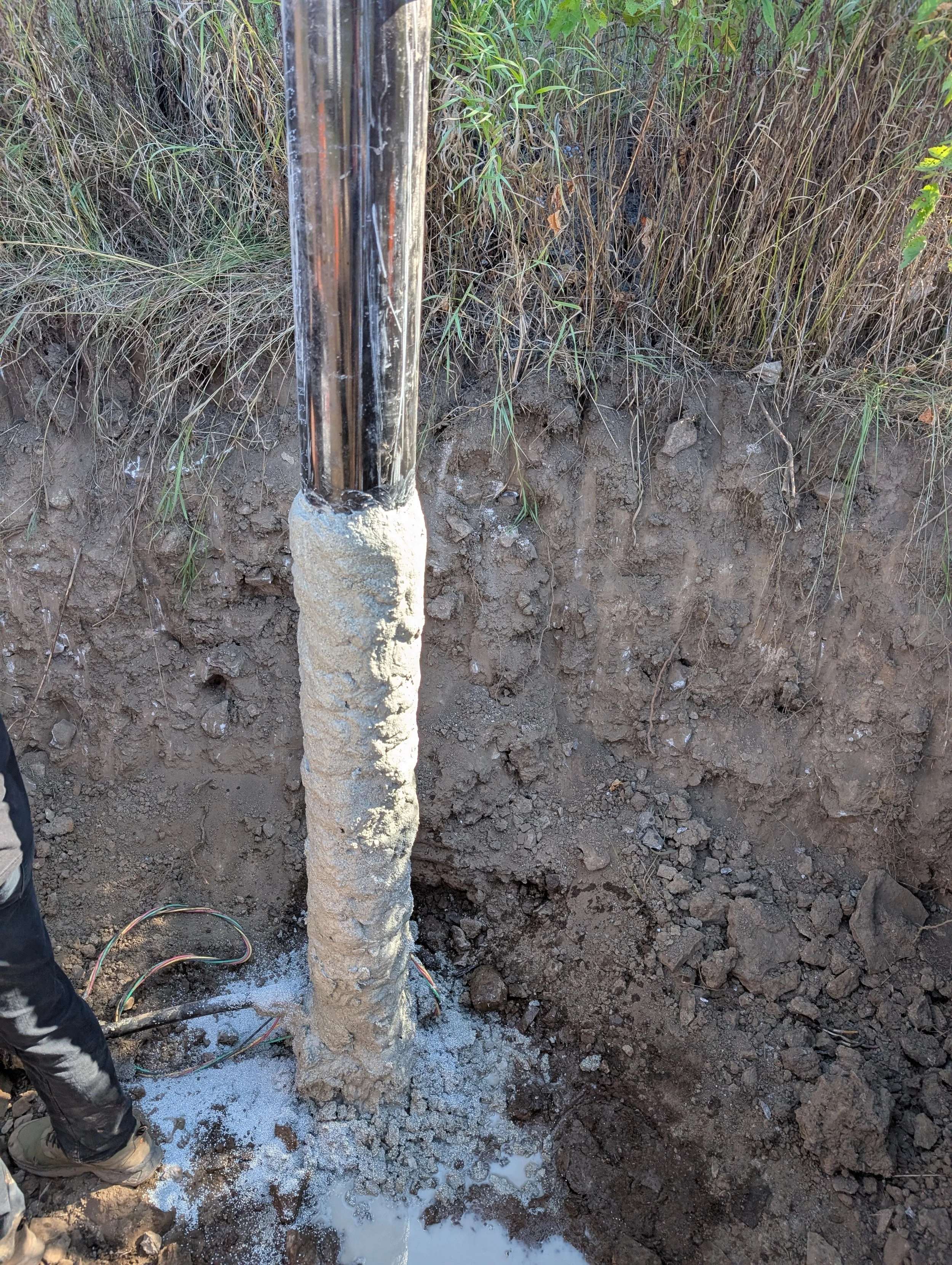 A vertical metal pipe surrounded by disturbed soil and muddy water, with green plants and grass in the background, and a person in black pants and shoes partially visible on the left side.