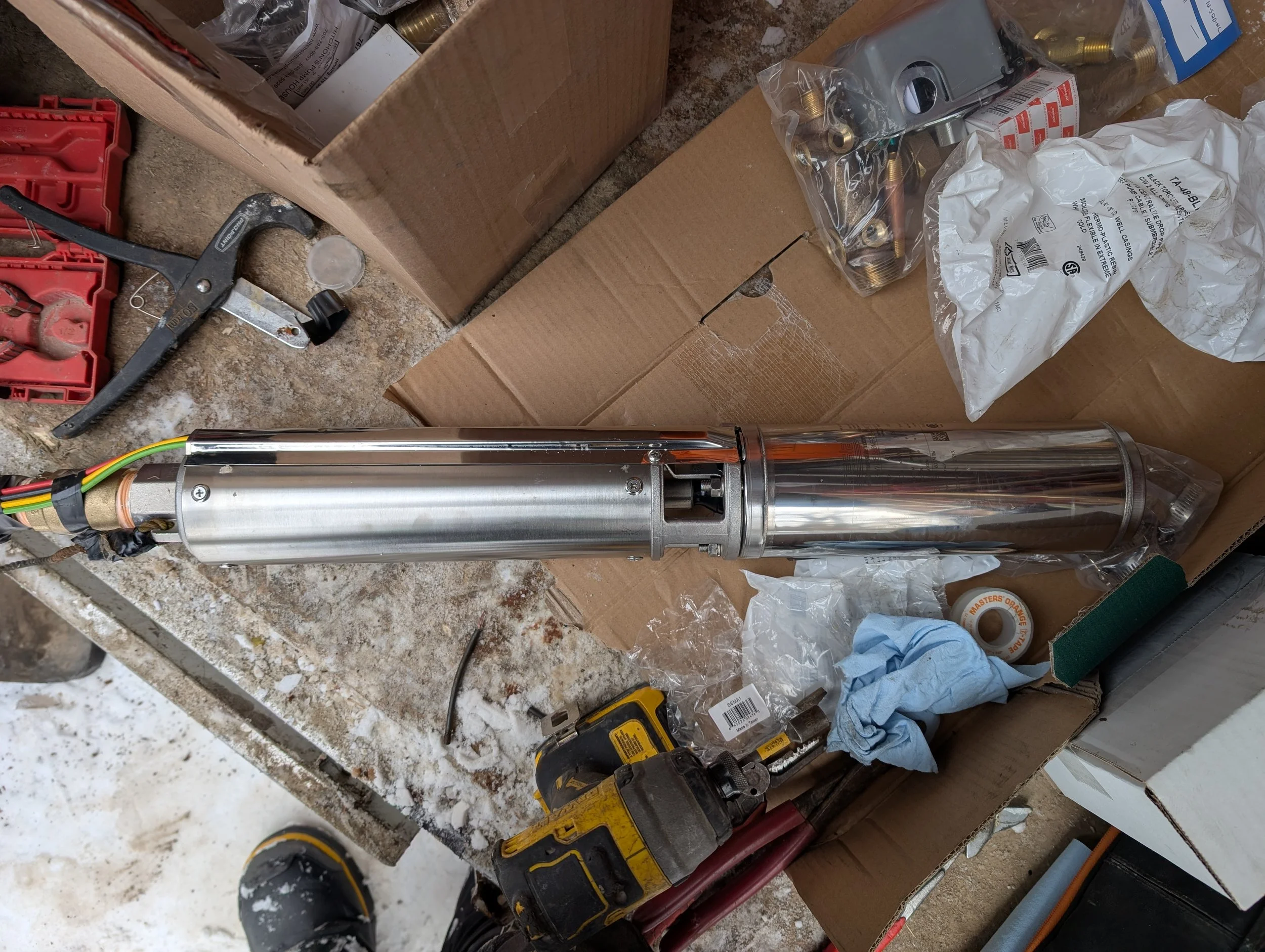 A cluttered workbench with tools and parts, including a large metal cylinder, power drill, screwdrivers, and various hardware items, on a dirt and dusty surface.
