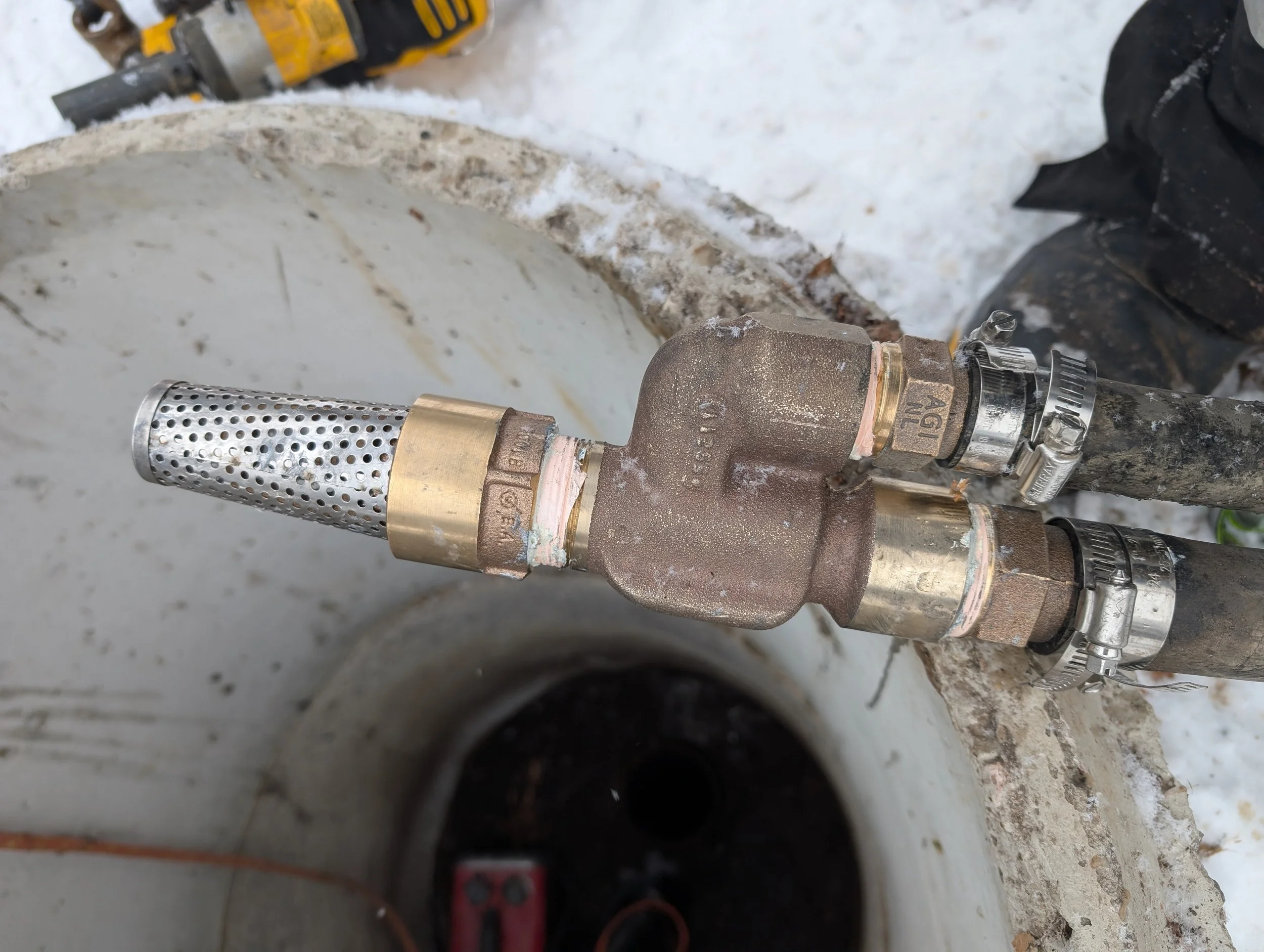 Close-up of a plumbing pipe fitting with a filter, surrounded by a rusty pipe, on a snowy ground, with a power tool in the background.