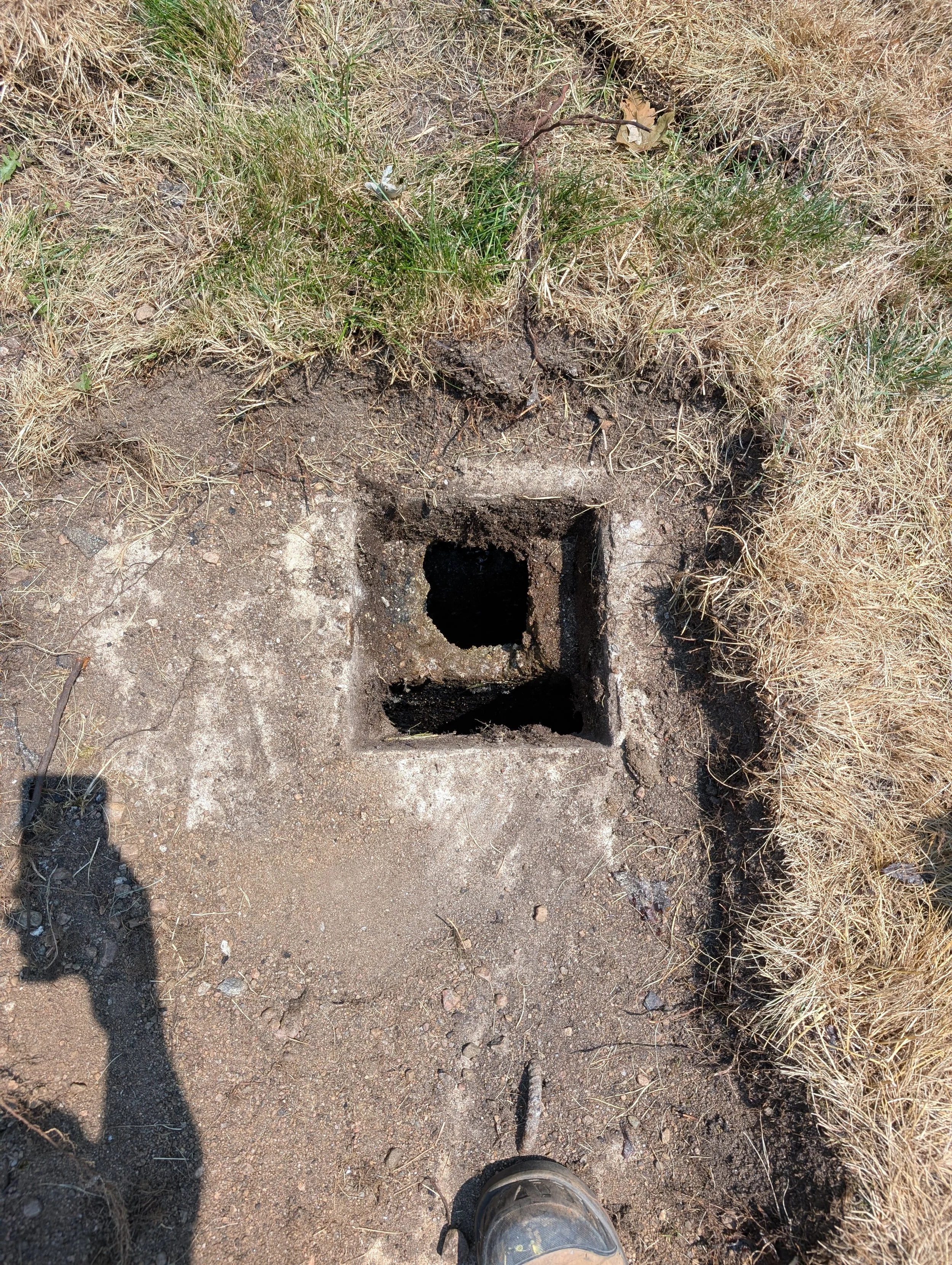 A hole in the ground surrounded by dry grass and dirt, with a person's shoe visible at the bottom of the image.