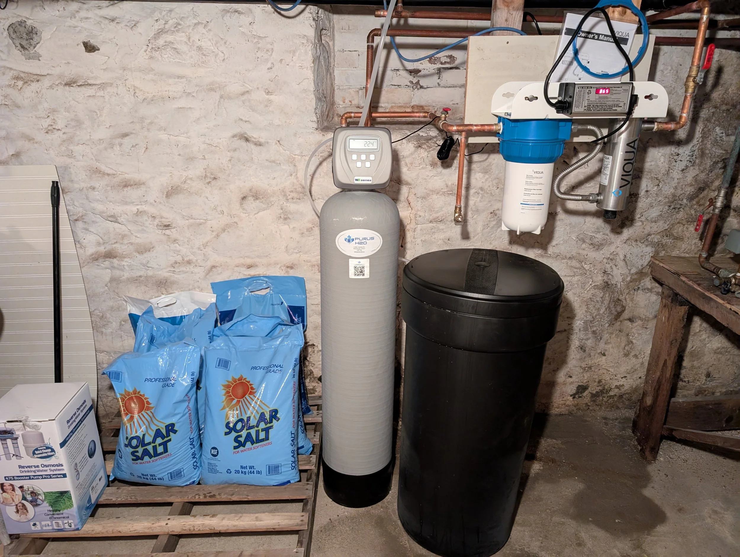 An indoor utility or basement room with exposed brick walls. A water filtration or treatment system is installed, including a large cylindrical tank labeled "Purus H2O" and a blue and white water filter unit. There are copper pipes, a digital control