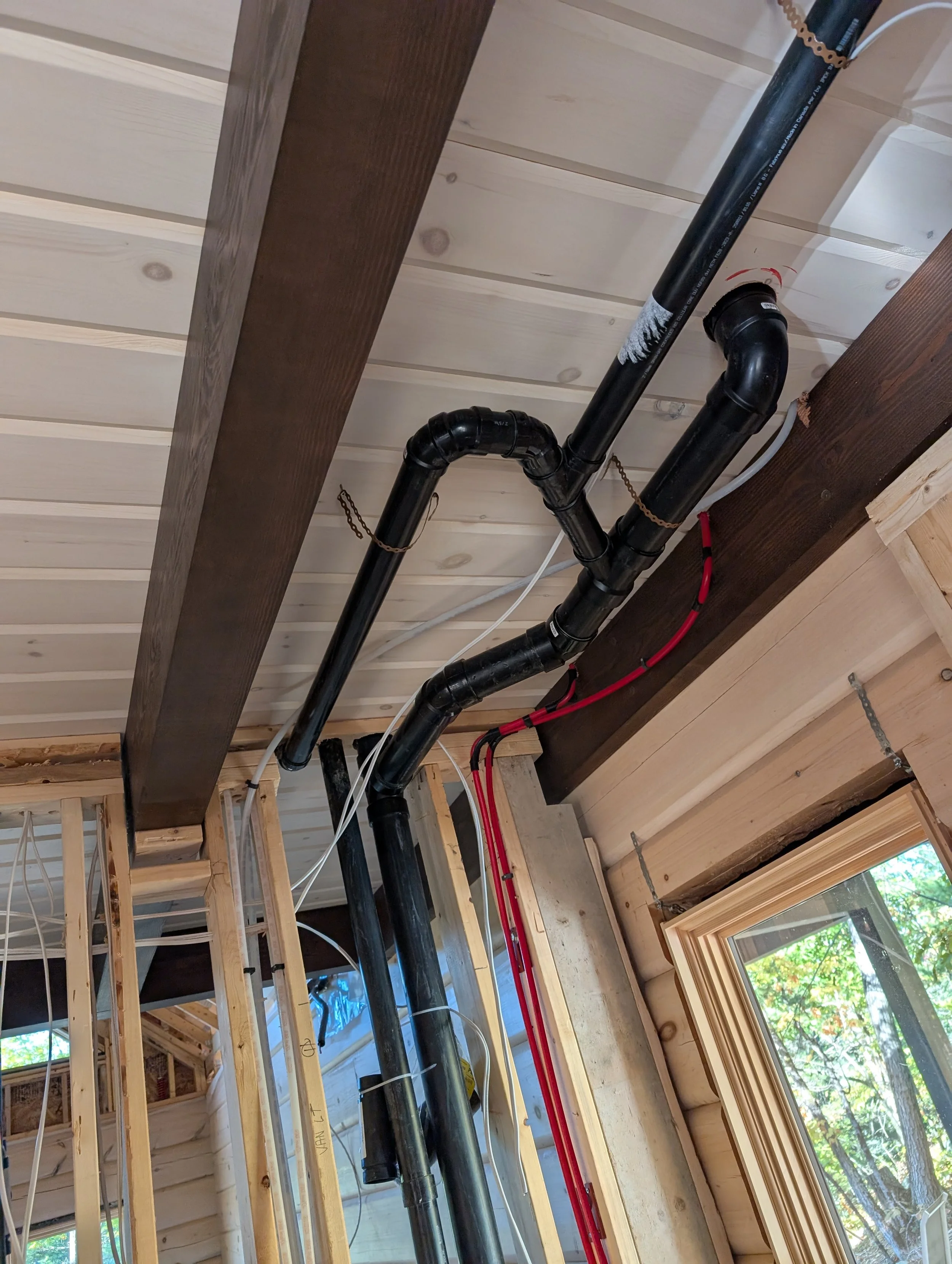 Interior of a building under construction with exposed wooden framing, black and red plumbing pipes, and a wooden window frame with a view of trees outside.
