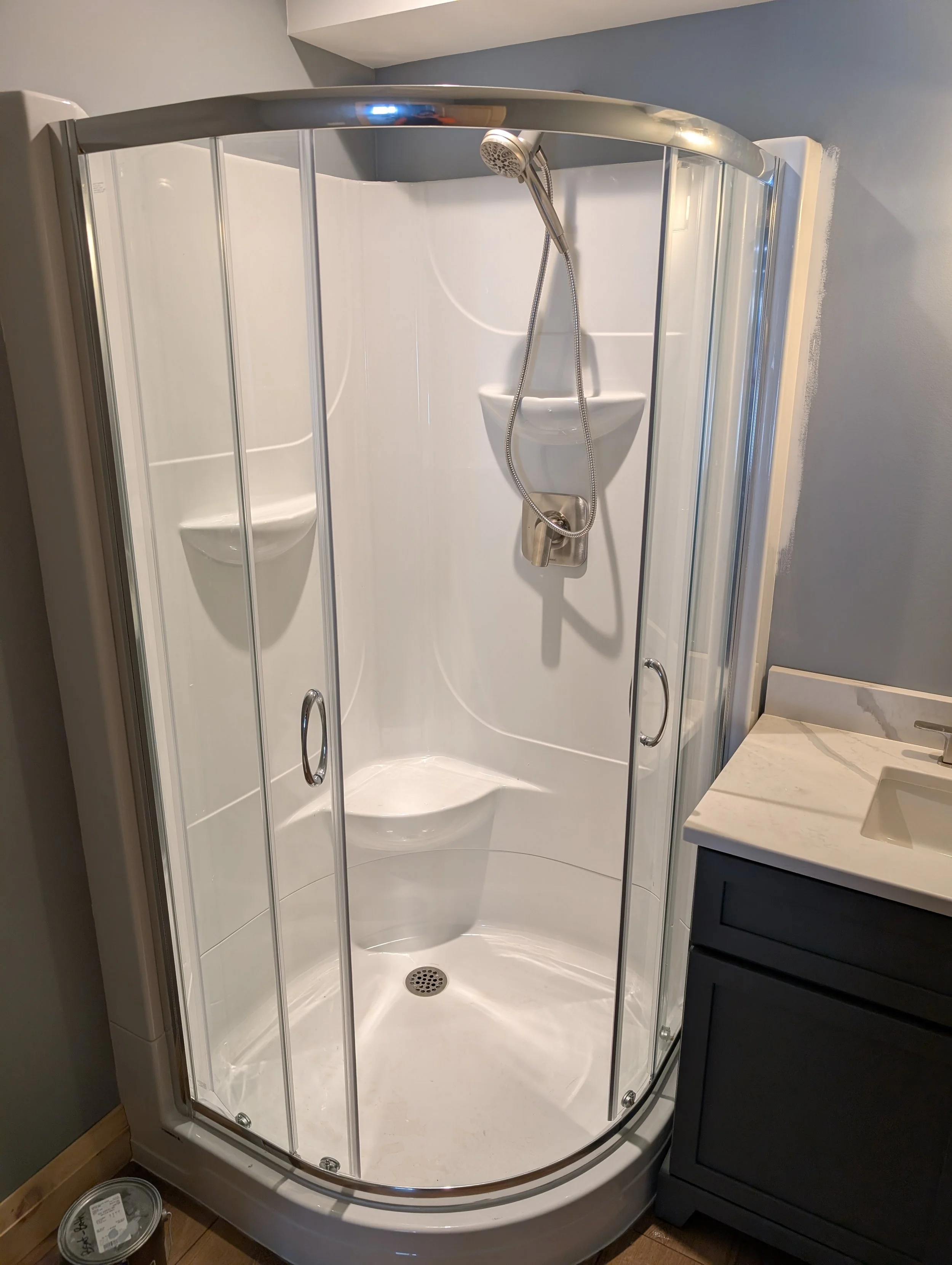 A corner shower stall with glass doors, a handheld shower head, a soap dish, and a small drain on the floor, next to a bathroom vanity with a marble countertop and a sink.