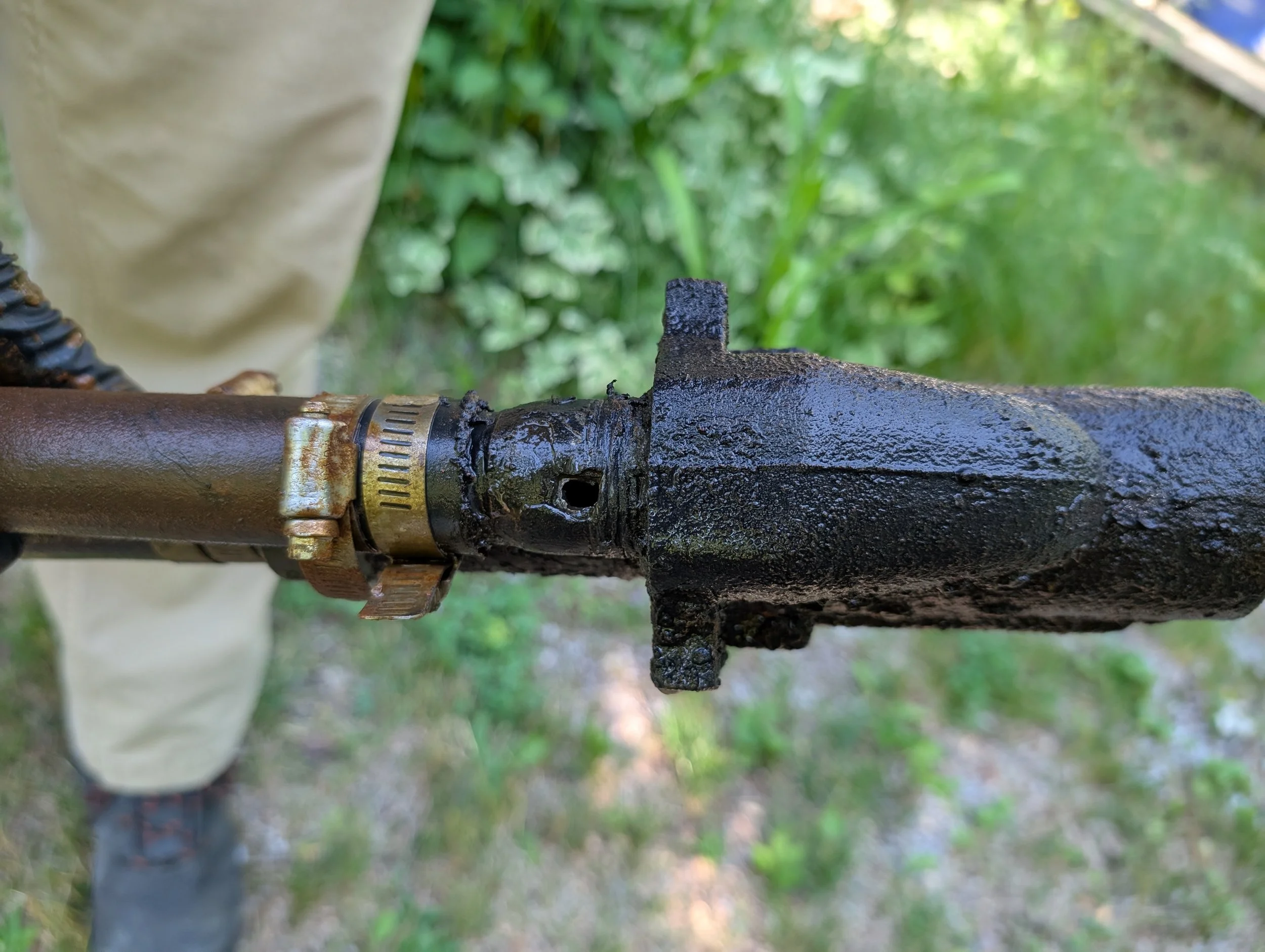 Close-up of a rusty, black pipe with a damaged and corroded fitting, with green foliage in the background.