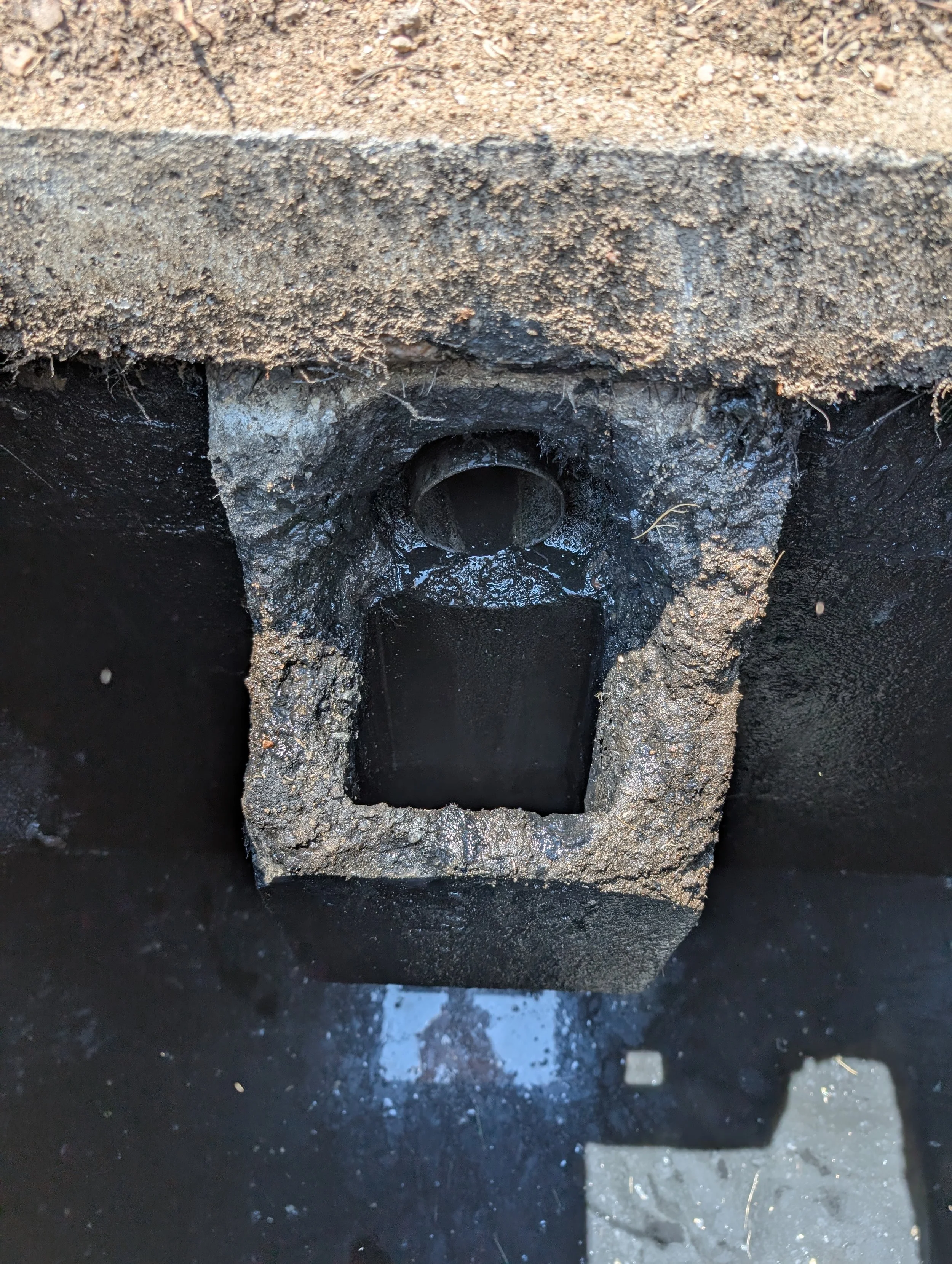 Close-up of a culvert with a black pipe embedded in a concrete and asphalt surface, surrounded by dirt and gravel.