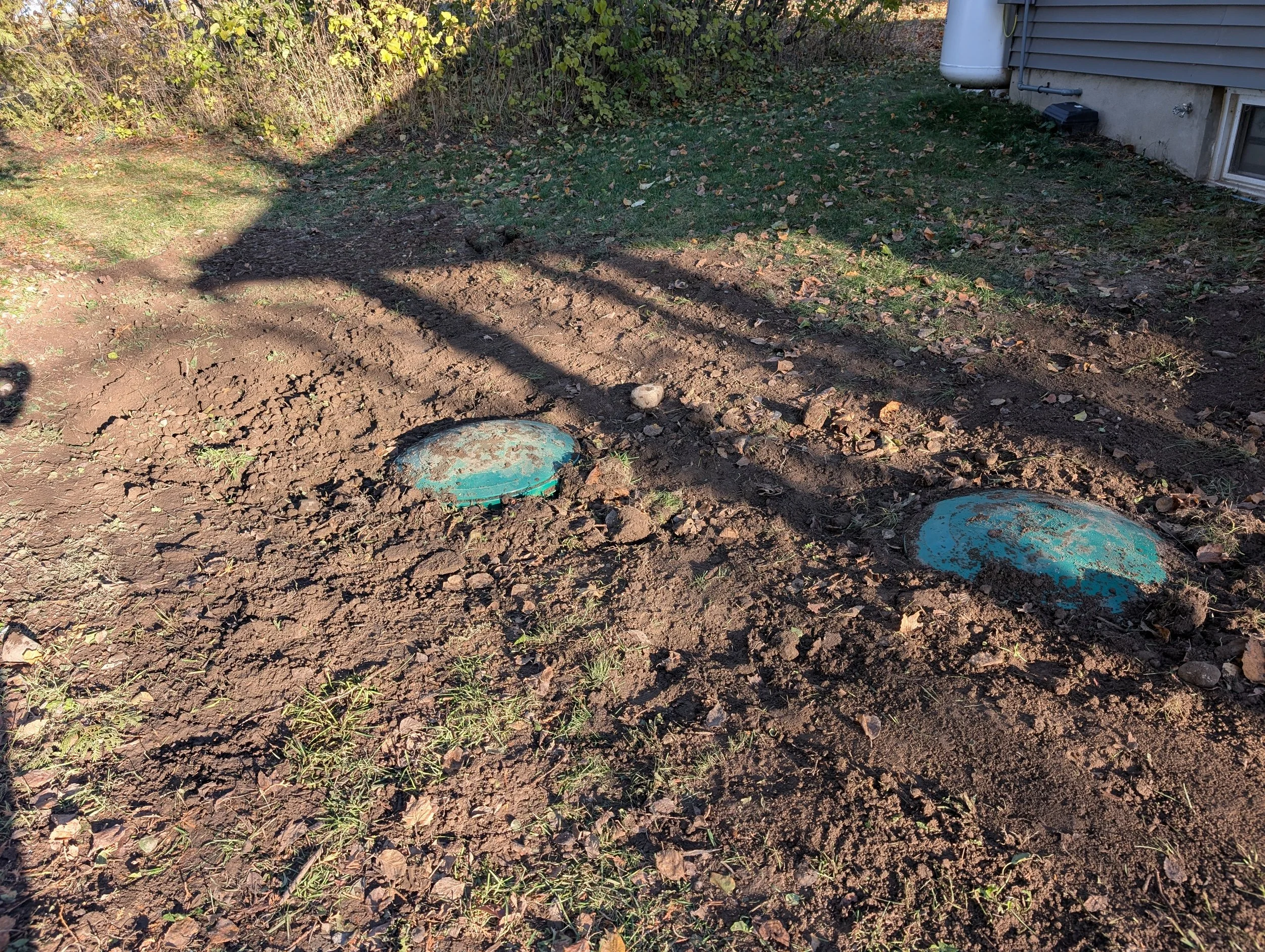 Exterior yard with dirt patch and two blue utility covers, adjacent to a house with gray siding and a small basement window, surrounded by fallen leaves and some green grass.