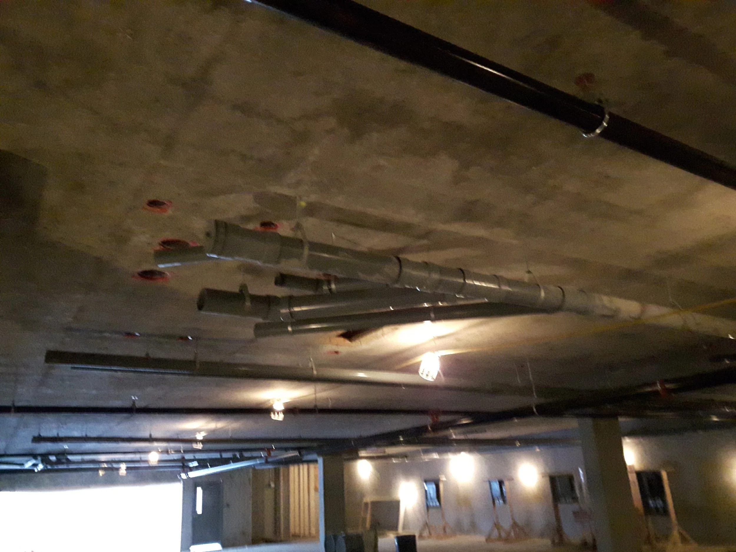 A dimly lit underground parking garage with concrete ceilings and multiple pipes running along the ceiling.