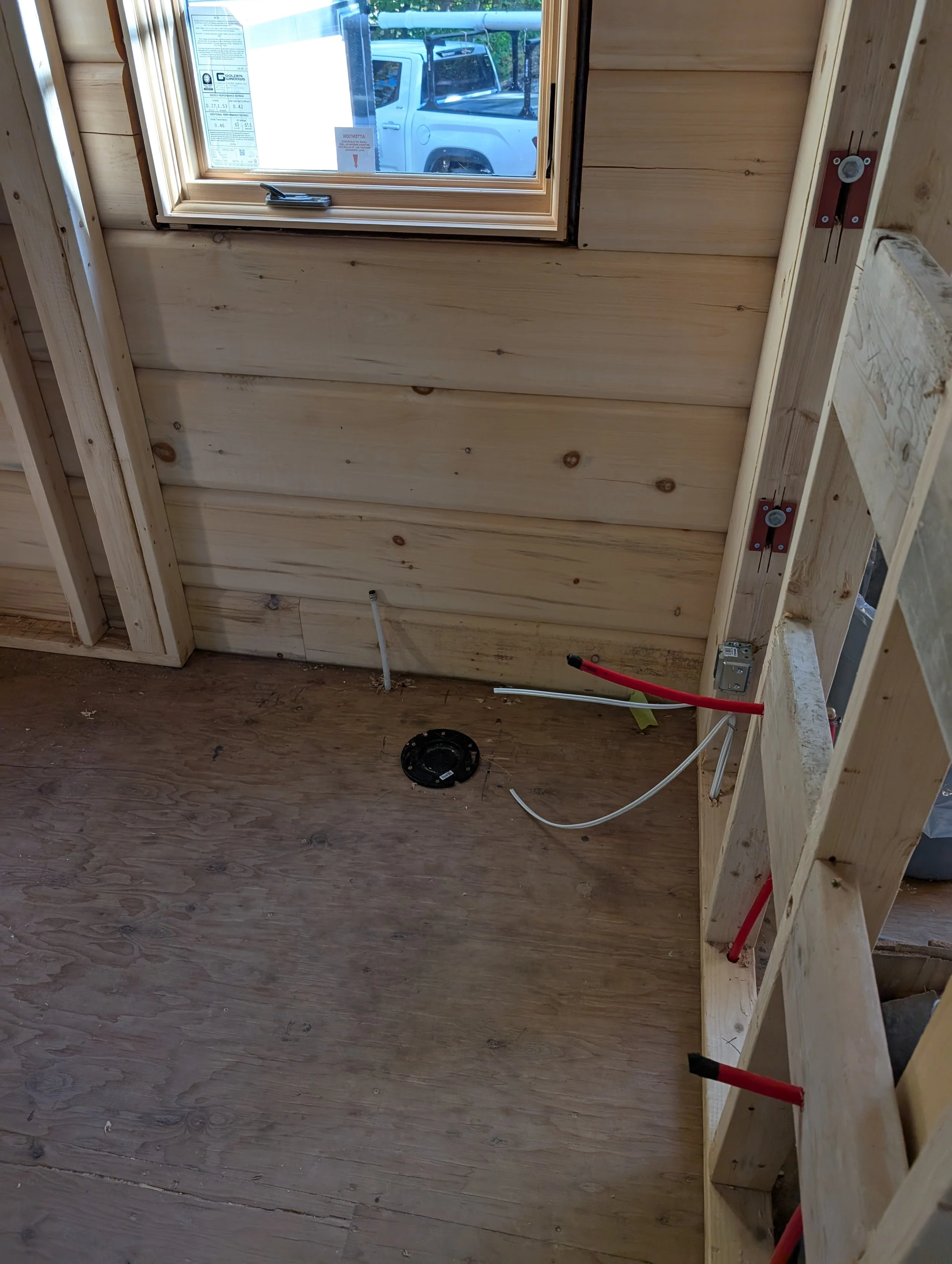 Interior view of a wooden wall under construction with a window, electrical outlet box, wires, and construction tools and materials visible.