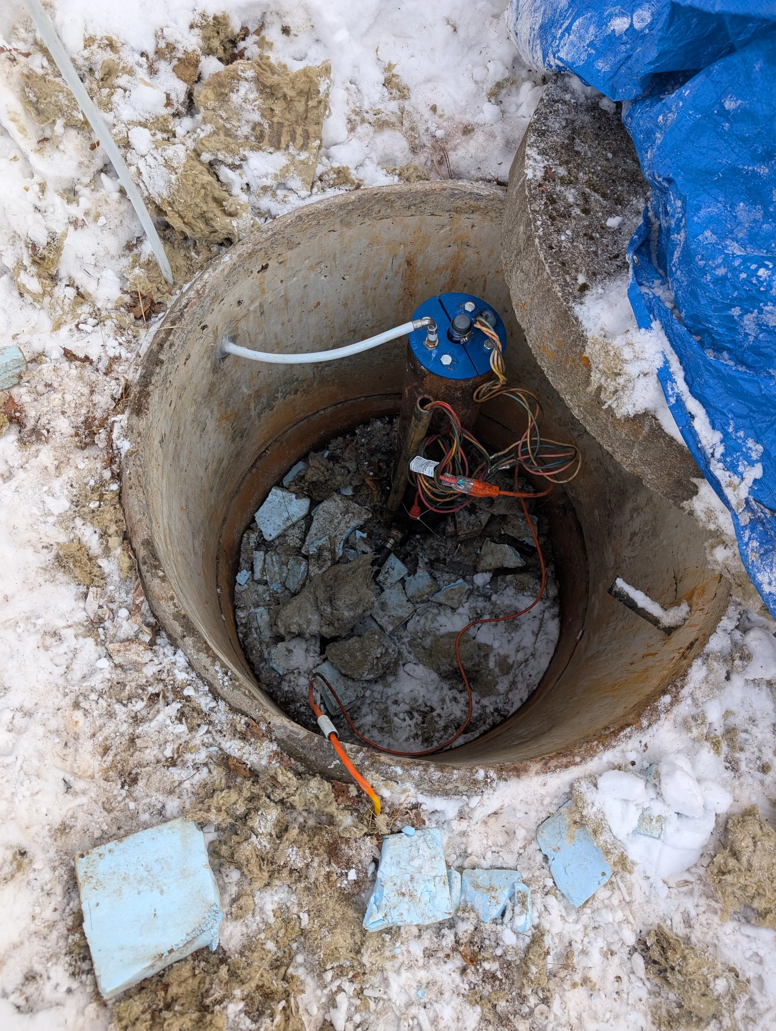 A construction site showing an open underground utility vault or manhole with electrical and communication cables, surrounded by snow and ice, with concrete debris and construction materials nearby.