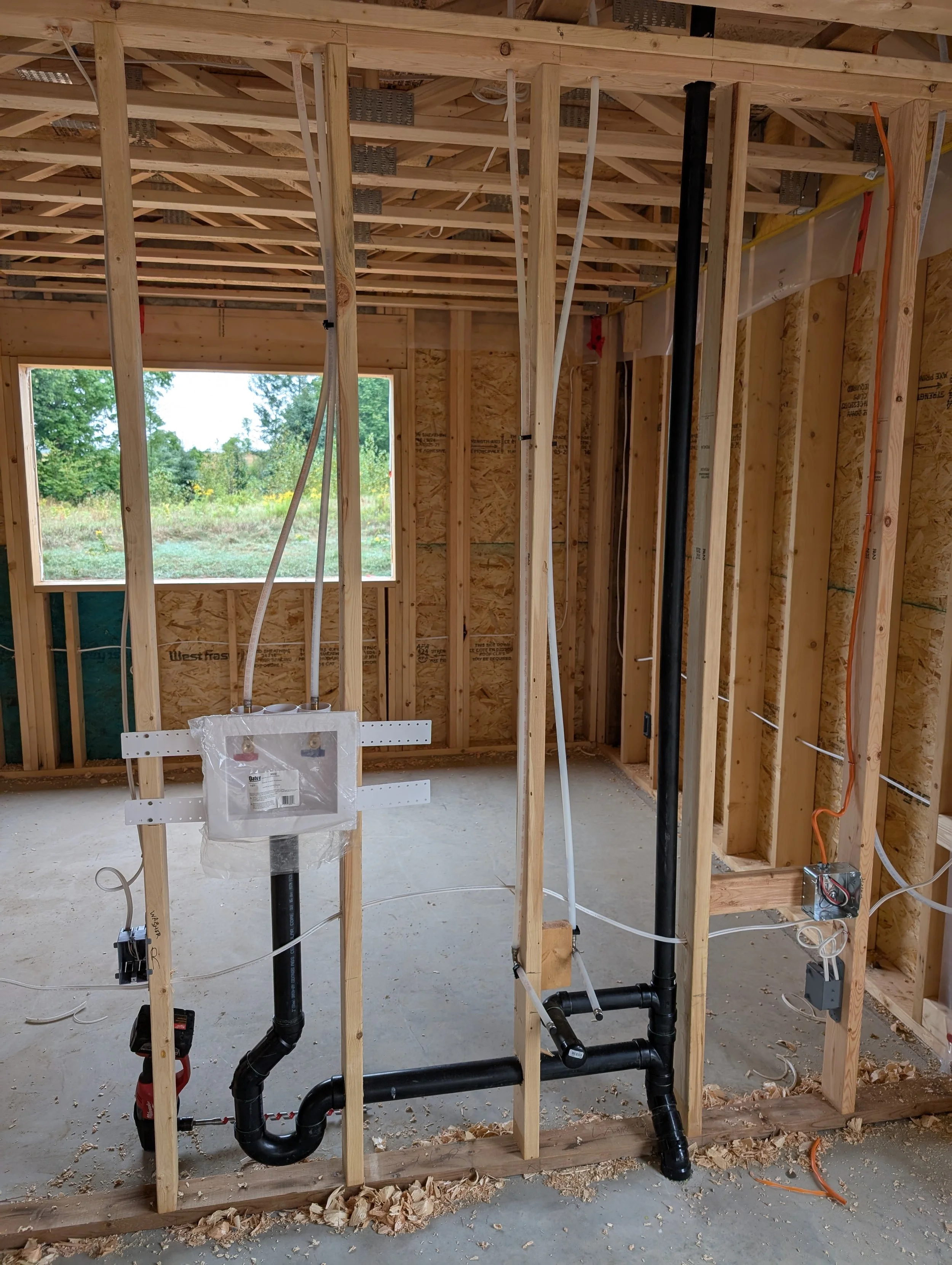 Interior of a house under construction with exposed wooden framing, plumbing pipes, and electrical wiring.