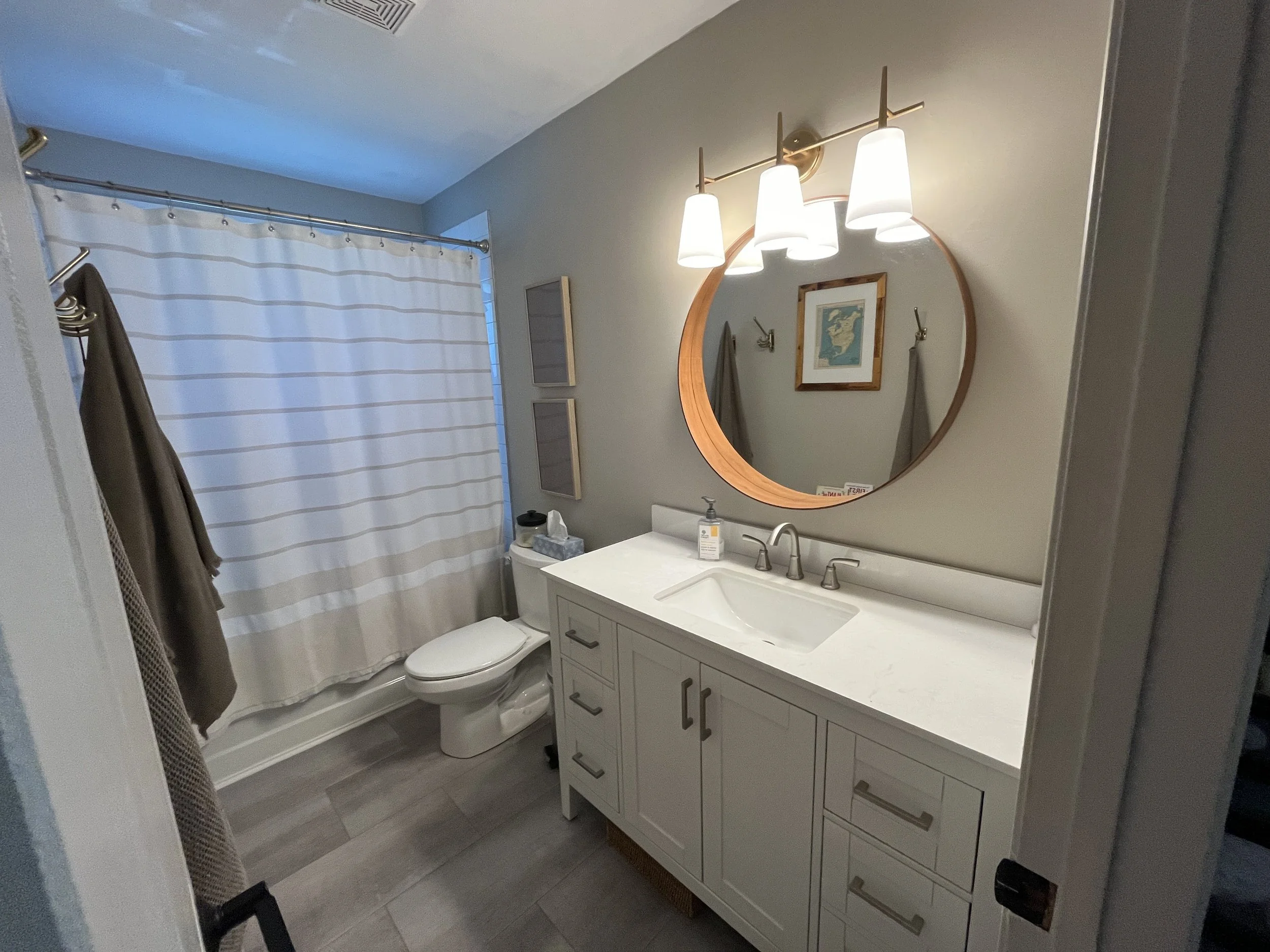 A modern bathroom with a white vanity, a round mirror with a wooden frame, a lighting fixture with five lamps, a shower with a striped curtain, a toilet, and decorative framed art on the wall.