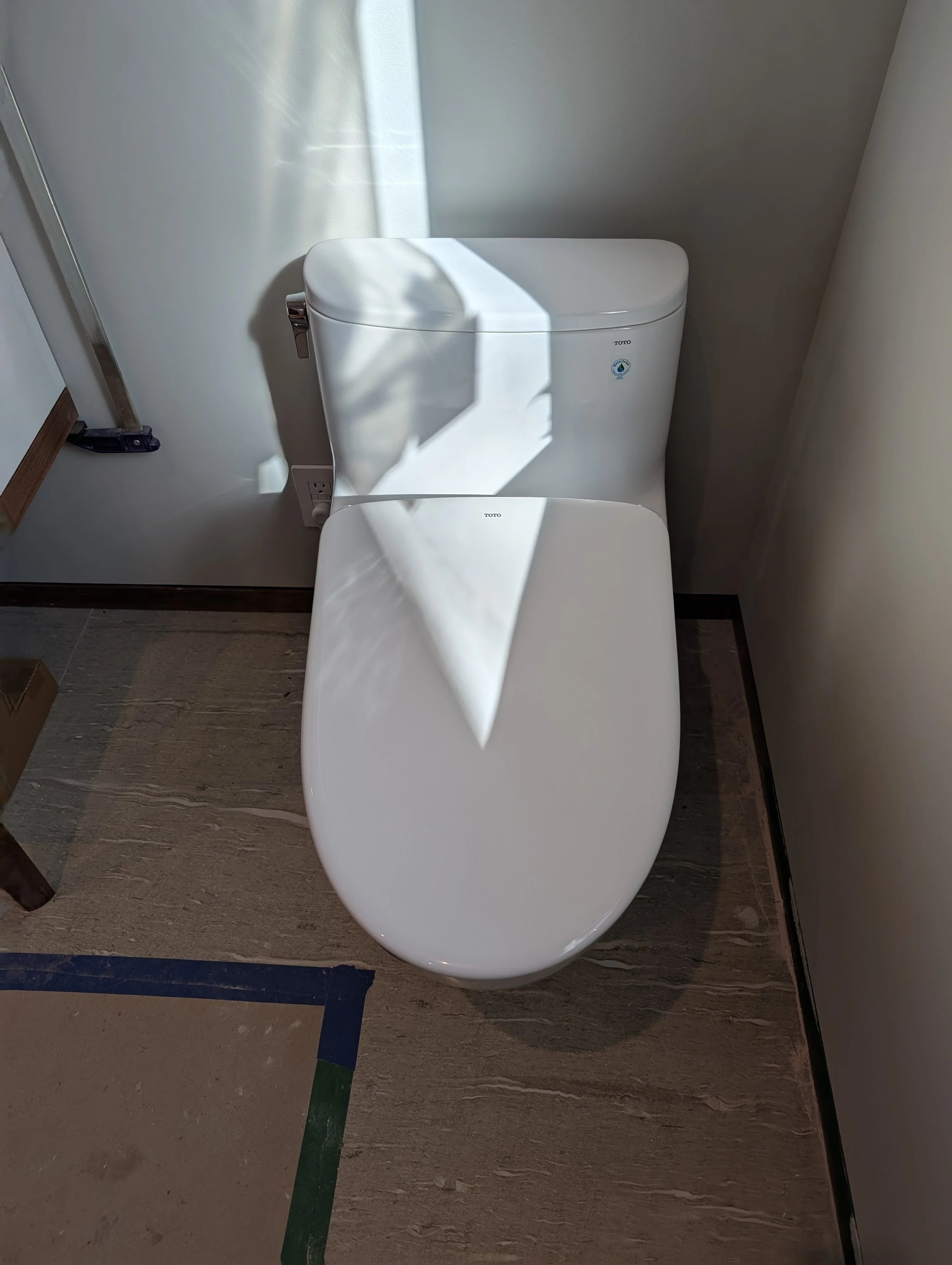 A modern, white, elongated toilet in a bathroom with beige and brown marble flooring. Sunlight casts shadows across the toilet and floor. Part of a wooden furniture piece and a power outlet are visible to the left.