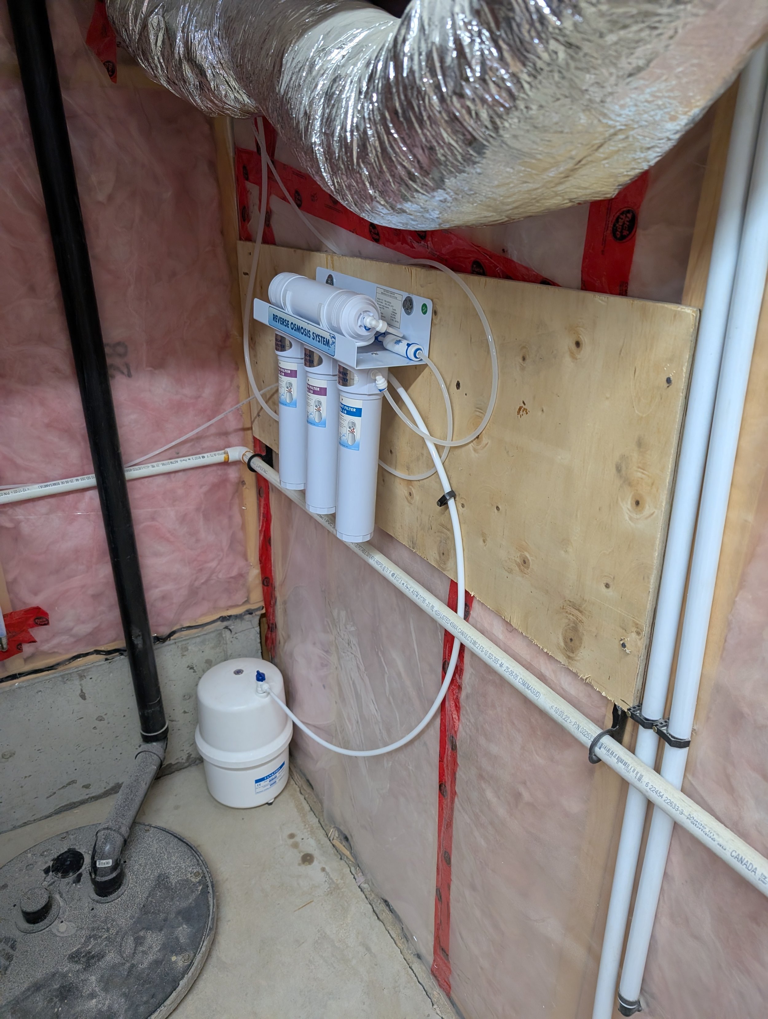 Under construction or renovation area showing a reverse osmosis water filtration system mounted on a wooden board, with pipes and a water filter. The area has exposed insulation and wiring, with a concrete floor and a gray pipe coming from the cold w