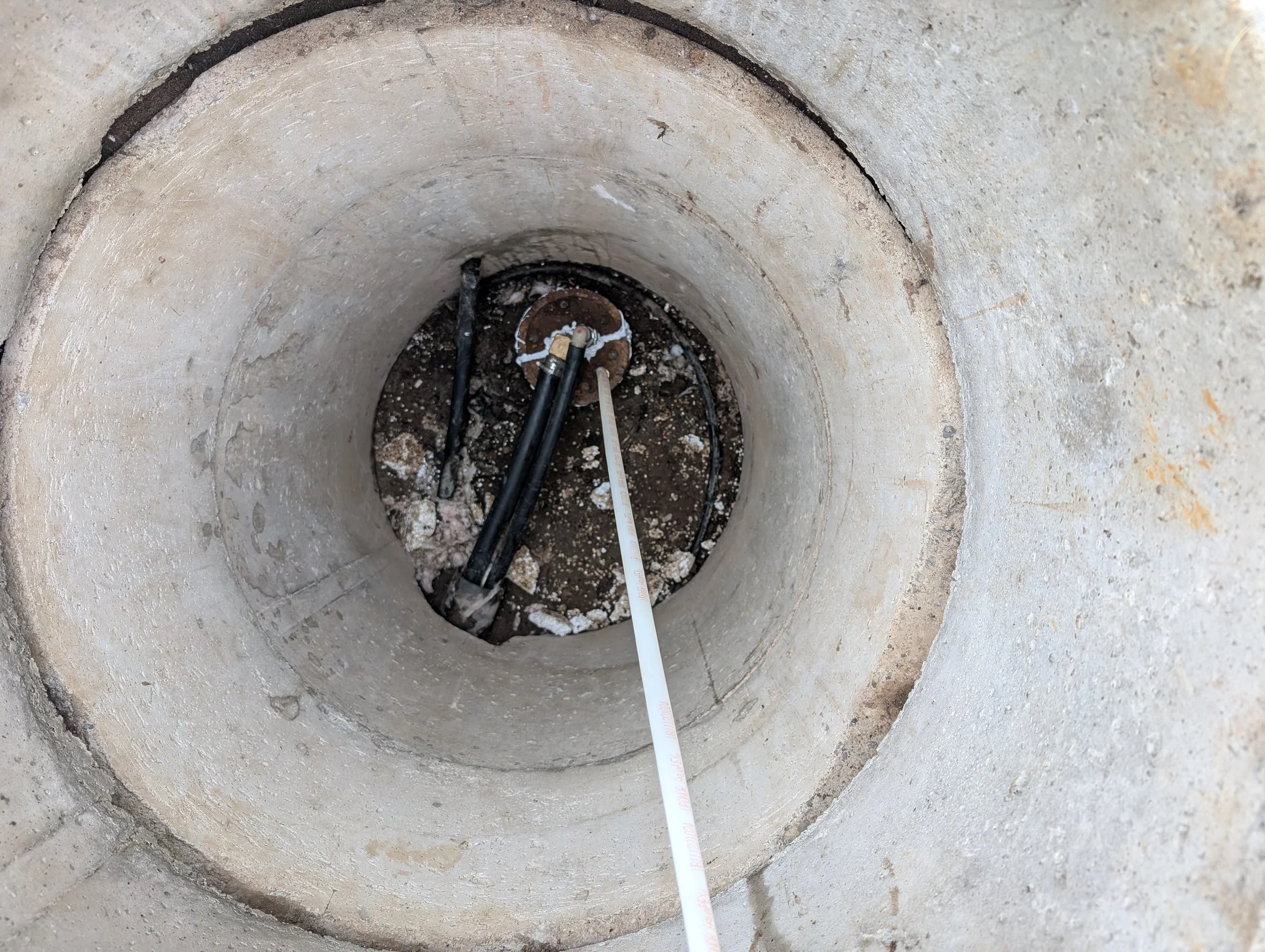 Looking down a deep, circular, concrete well with a white measuring tape inside. The well's interior and surrounding rim appear rough and worn.